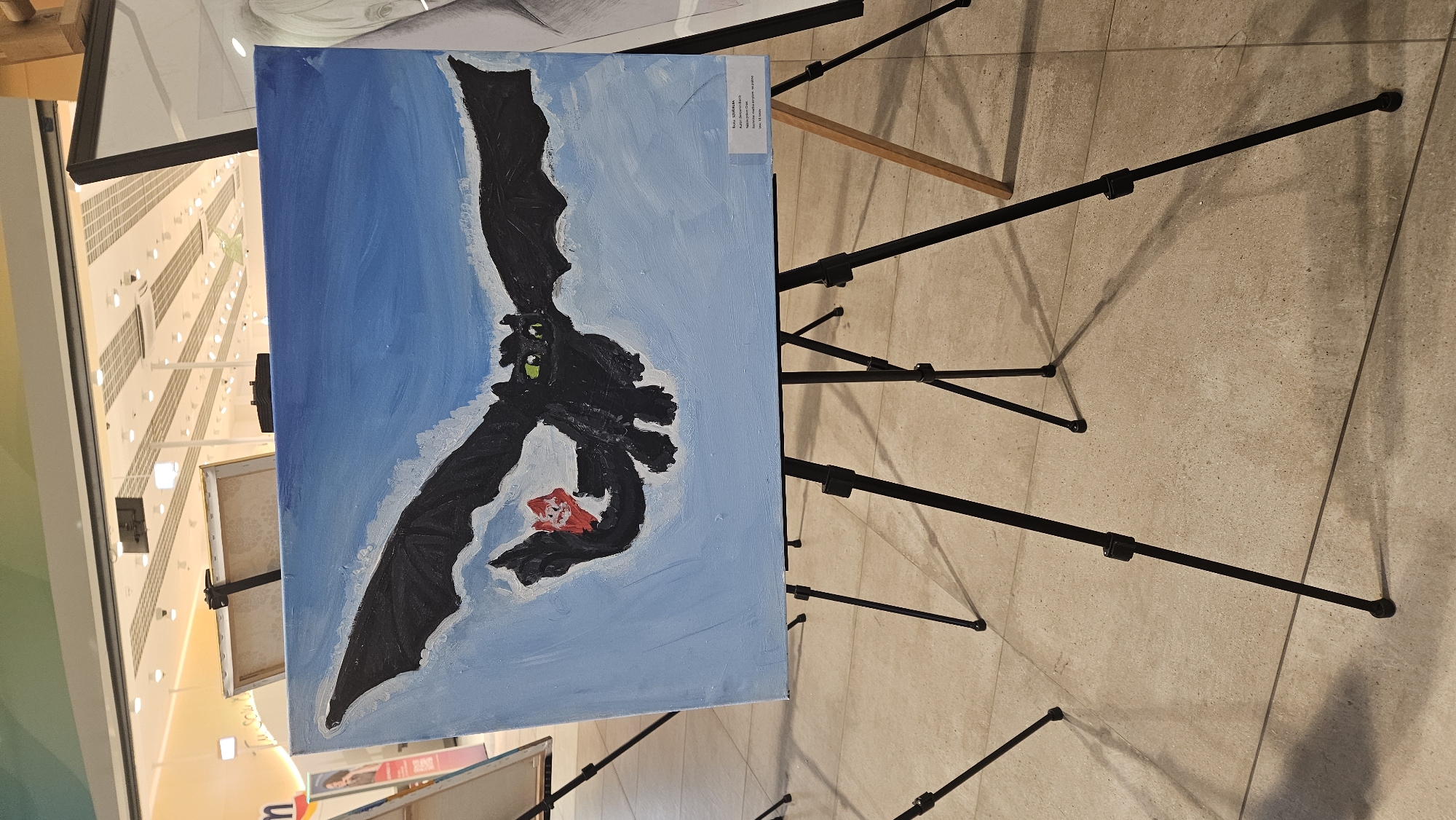 The image shows a painting of Toothless, the dragon from "How to Train Your Dragon," flying against a light blue sky with white clouds. The dragon is black with green eyes and has a red fish in its mouth. The painting is on a canvas and is displayed on a black tripod easel. A small white label is attached to the bottom right of the canvas, but the text is illegible. The painting is located indoors, possibly in a public space like a mall or exhibition hall, suggested by the tiled floor and the background with artificial lighting and ceiling structures. There is a glimpse of another artwork to the right of the main painting. The overall scene suggests an art exhibition or a display of student artwork. The location of Prešov, Slovakia, is not visually discernible from the image itself.