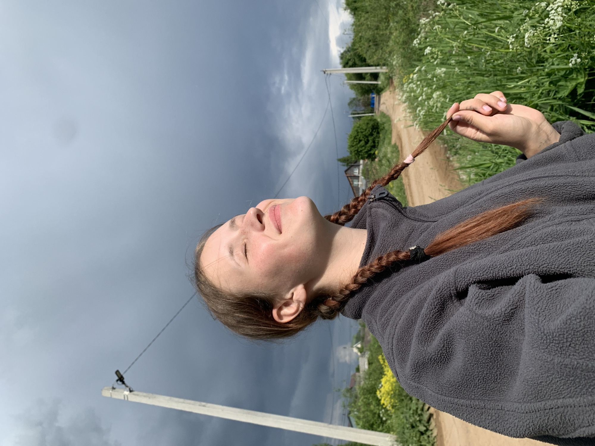 A woman with brown hair styled into two braids stands outside on a dirt road, looking up towards a stormy sky. She wears a grey fleece jacket and holds the end of one of her braids in her hand. The surrounding area features green foliage and a few buildings in the distance. Utility poles line the roadside. The picture was taken in Фоки, Russia.