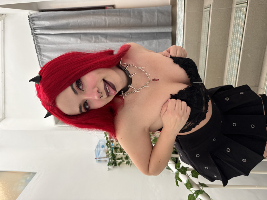 The image displays a young woman with vibrant red hair, presented in a portrait orientation that has been rotated 90 degrees clockwise. She has fair skin and striking facial features, including dark eye makeup with winged eyeliner. She wears several facial piercings: a septum piercing, a philtrum piercing, and a tongue piercing, which is visible as her tongue is slightly extended. Two small, black, horn-like accessories are positioned within her red hair, just above her forehead. Her expression is a playful smile, directed towards the viewer.
Her attire consists of a black, spiked choker around her neck. This choker is connected by a chain to a second, more delicate chain necklace that features small, spike-like elements and a distinct red, coffin-shaped pendant. She is wearing a black lace bralette or bustier top. Her right hand is visible, gently holding the fabric of her top, while her left hand is partially obscured, also seemingly adjusting her top. She is also wearing a black pleated mini skirt adorned with numerous silver-colored metal grommets or eyelets.
The background setting appears to be indoors. To her left (which would be the top if the image were upright), a grey curtain or blind is visible against a light-colored wall. To her right (the bottom when upright), a white staircase with railings can be seen, partially covered by green foliage or artificial vines. A glimpse of what might be a birdcage or a similar decorative object is also visible in the mid-ground. The lighting suggests a bright, possibly daylight, indoor environment.
