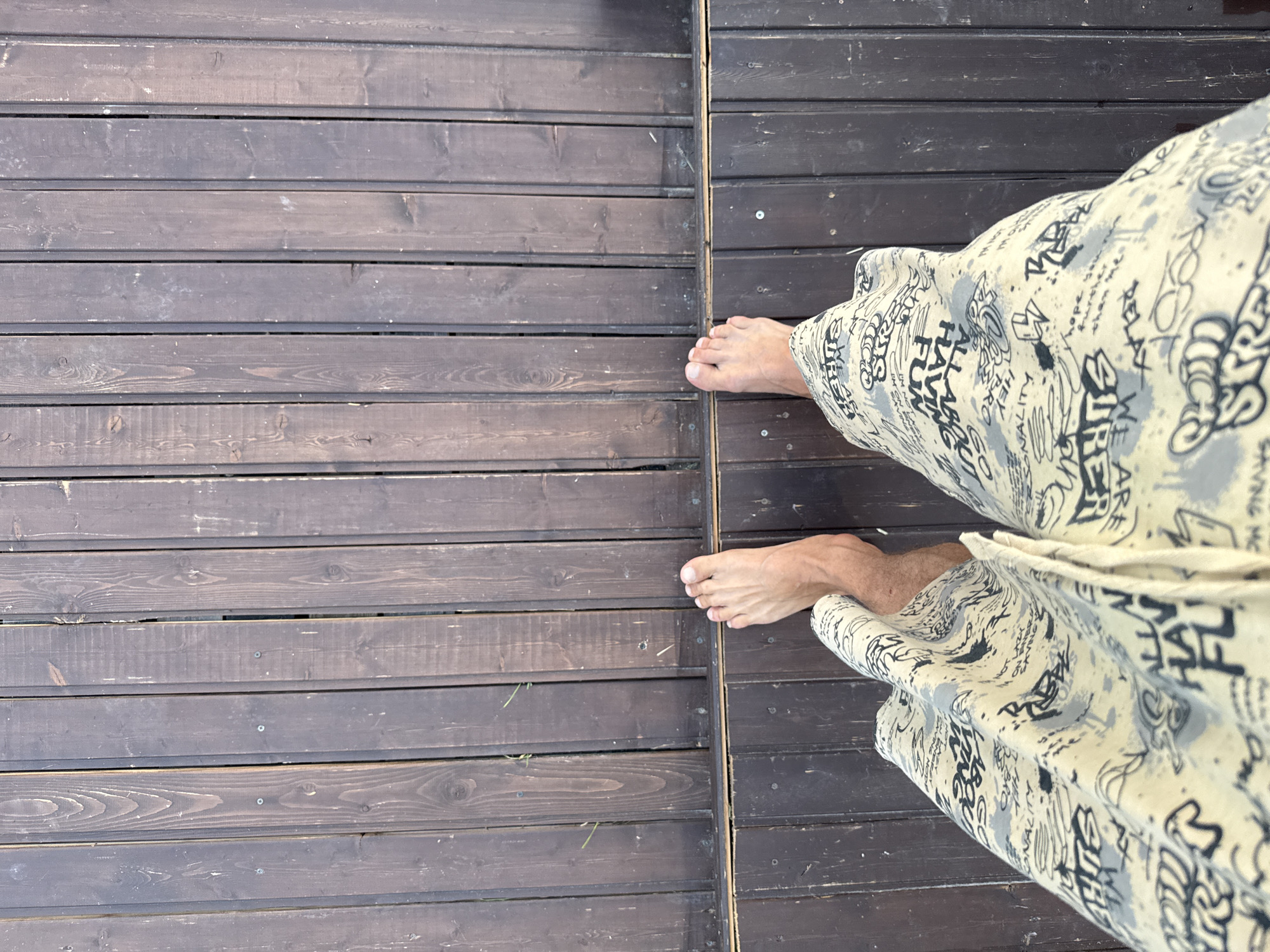 A first-person perspective shot shows a person's bare feet and patterned shorts standing on a wooden deck. The deck is made of dark brown planks running vertically, with some grass growing between the planks. The shorts have a light yellow base with black graffiti-style text and designs. One foot is positioned slightly in front of the other, with the toes of the front foot visible. The image is taken from a high angle looking down. The location is not specified in the image.
