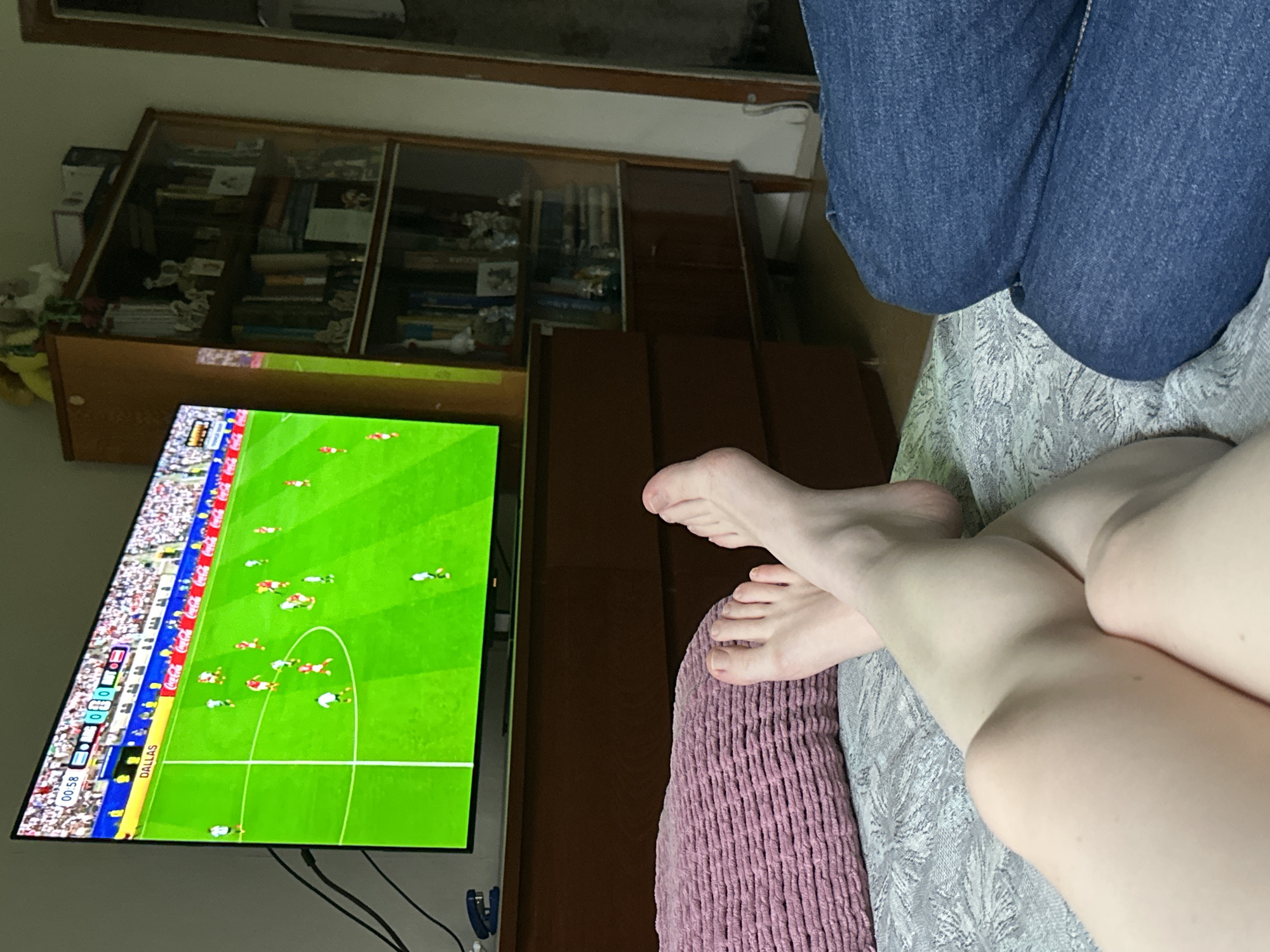 A person's legs and feet are visible in the foreground, resting on a bed. The person is wearing blue jeans. In the background, a television screen displays a soccer match. The television is placed on a wooden dresser. To the right of the dresser, a wooden bookshelf filled with books and decorative items is visible. The room appears to be a domestic setting. The location is not specified as Moscow, Russia.