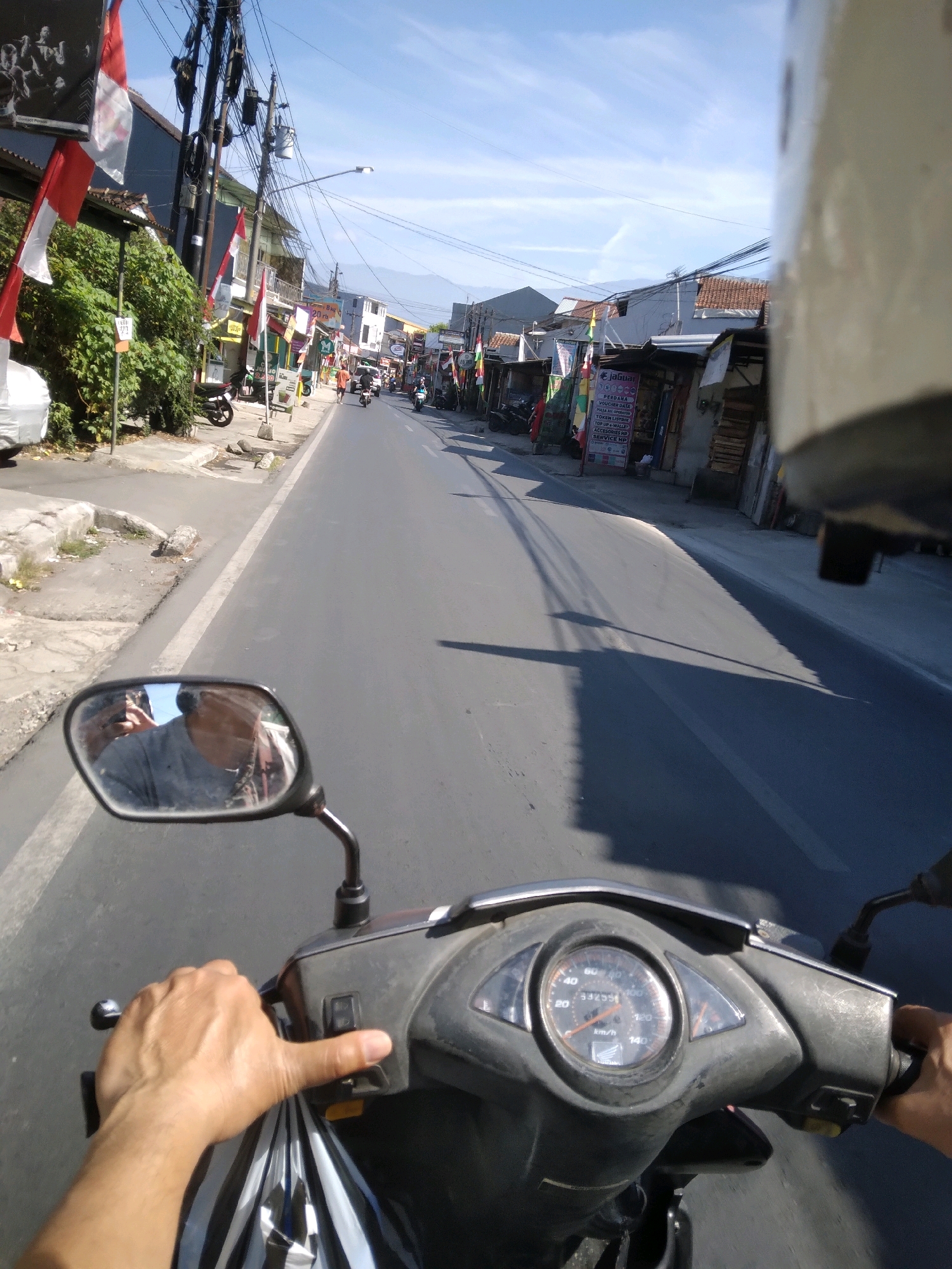 The image shows a first-person perspective of someone riding a motorcycle on a paved road in Grendeng, Indonesia. The rider's hands are visible, one on the handlebar and the other holding a bag. The motorcycle's dashboard, including the speedometer, is in the foreground. The road is lined with buildings, shops, utility poles with many wires, and trees. Indonesian flags are displayed in several locations along the street. The sky is blue with some clouds. In the distance, mountains are visible. The scene appears to be during daylight hours.