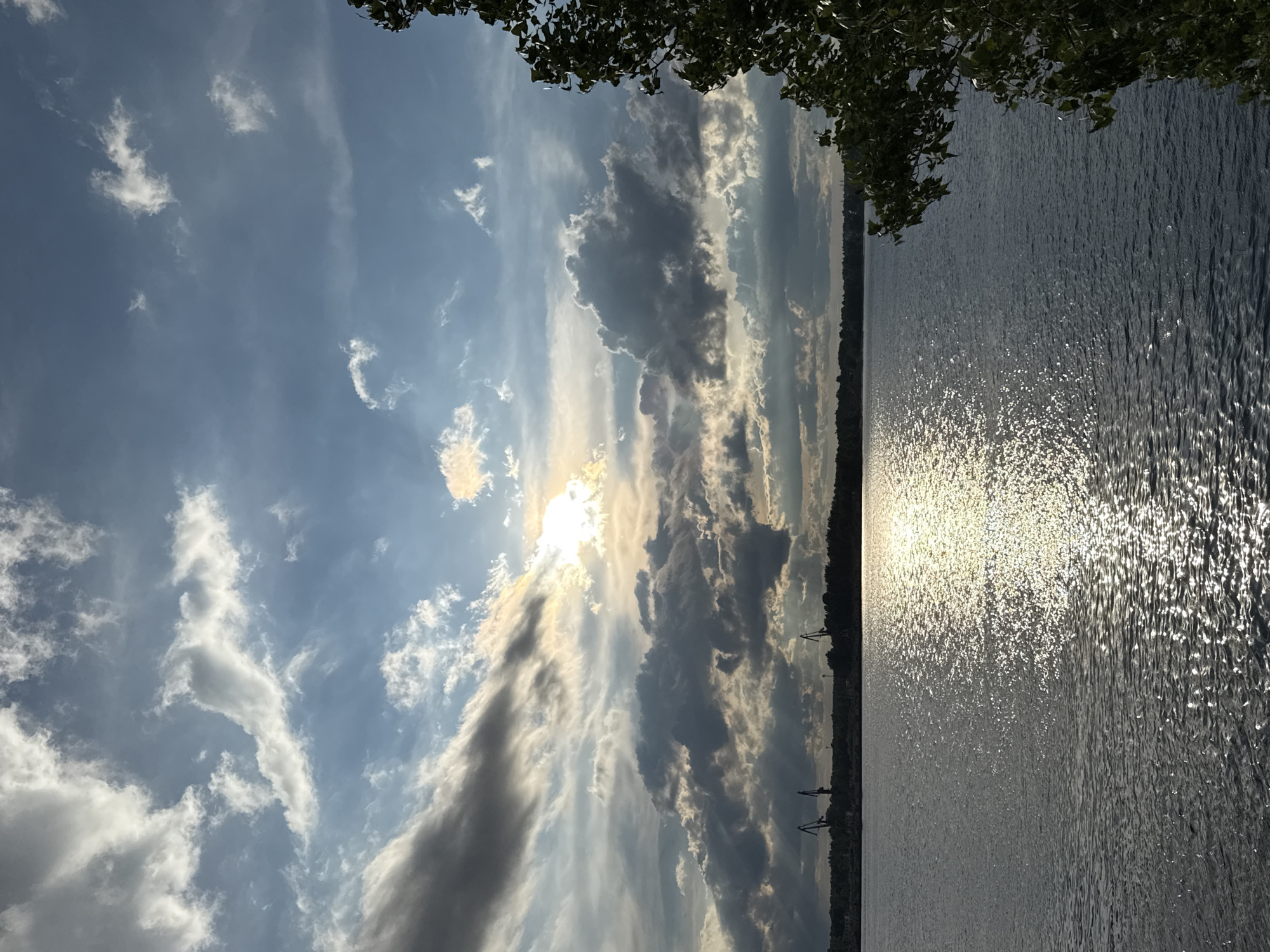 The image depicts a sunny day over a body of water. The sun is bright, reflecting off the water's surface. Clouds fill the sky, some dark and heavy, others lighter and wispy. Trees are visible along the shoreline. The location is the city of Chaikovsky, Russia, with the river Kama visible. There are cranes near the riverside.