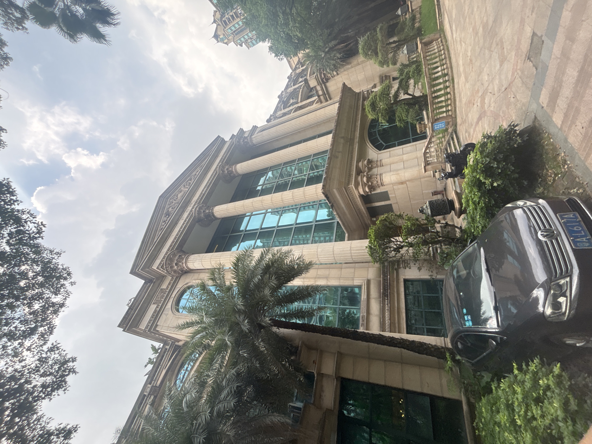 This is a photograph of a large, classical-style building with ornate columns and large, reflective blue windows, likely located in the city of Nanhai District, China. The building is surrounded by lush green foliage, including palm trees. In the foreground, a dark-colored sedan is parked, partially obscured by the vegetation. A portion of a license plate, reading "AL617A", is visible on the car. To the right of the car and slightly further back, a motorcycle is parked next to a stone railing. The sky above is overcast with some clouds visible. The overall impression is one of a well-maintained, possibly upscale residential or commercial property.