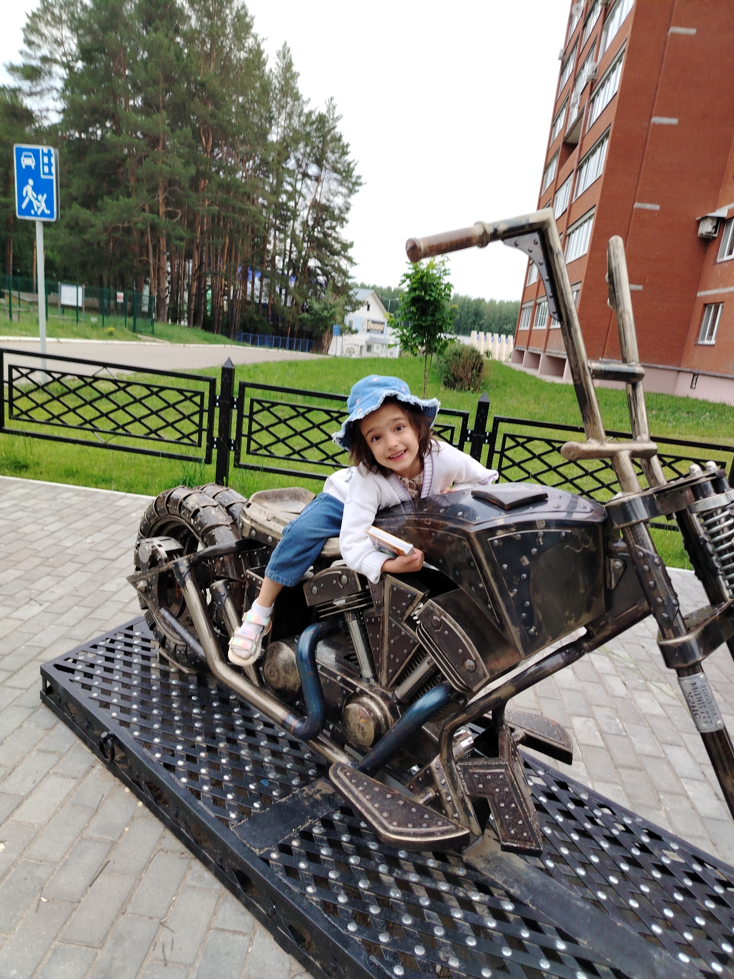 A young girl wearing a blue hat and a white sweater sits on a metal sculpture of a motorcycle. She is holding a small book in her right hand and smiling at the camera. Her left leg is extended, and she is wearing sandals. The motorcycle sculpture is dark and metallic, with visible engine details and a large tire. It is mounted on a black metal platform with a diamond plate pattern.
The background shows a paved walkway, a black metal fence with a diamond pattern, a grassy area, and a tall, red brick building. Behind the grass, there are numerous tall pine trees. To the left of the motorcycle, a blue and white traffic sign is visible, depicting a car and two pedestrians. The setting appears to be outdoors, likely in a park or public area in Chaikovsky, Russia. The lighting suggests it is daytime, with an overcast sky. The girl's expression conveys happiness and engagement with the camera. The overall scene is cheerful and portrays a child enjoying a playful moment with a unique art installation.