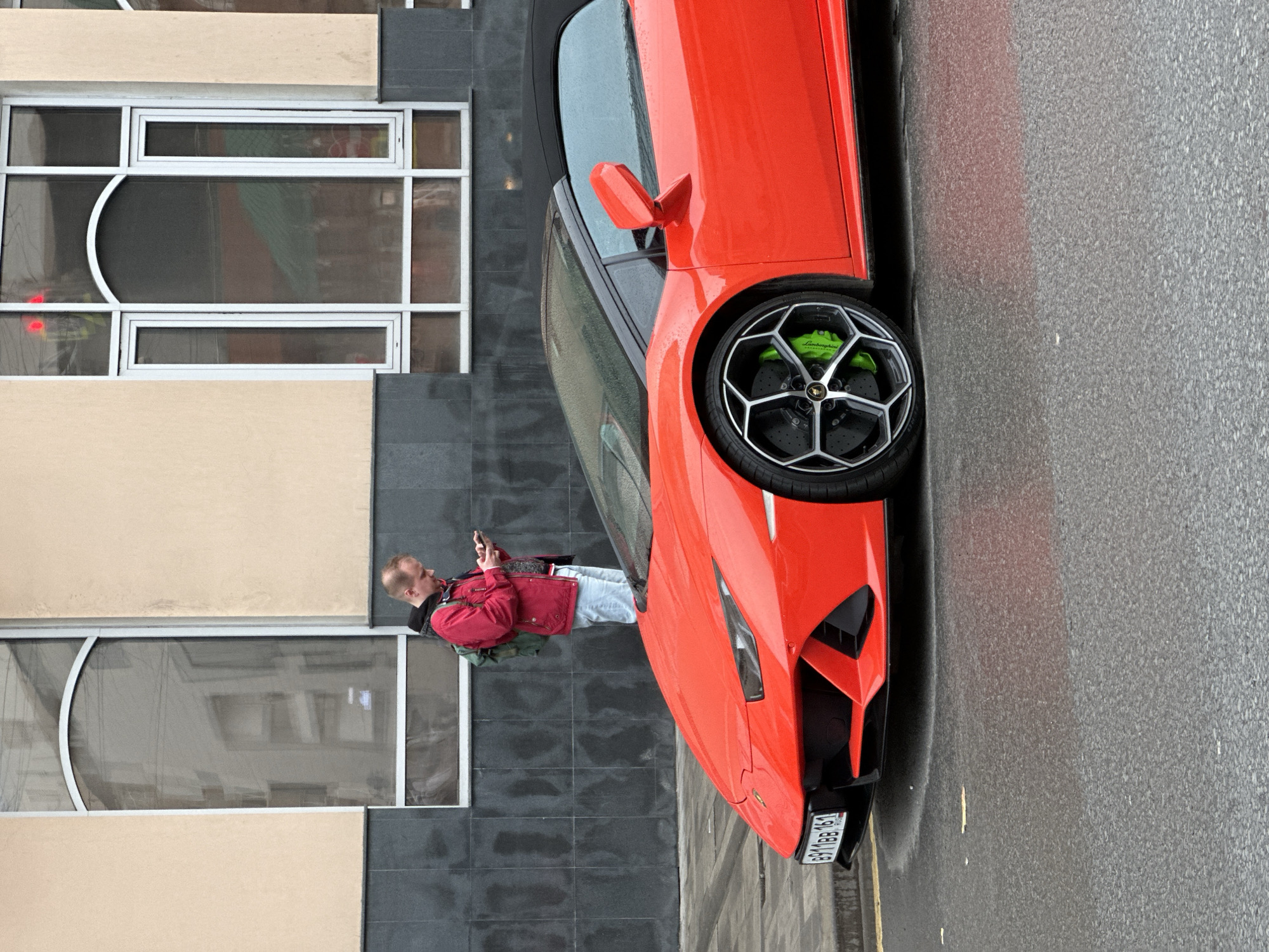 A red Lamborghini convertible is parked on an asphalt street in Rostov-on-Don, Russia. A man wearing a red jacket and light-colored jeans is standing next to the car, looking at his phone. The car is parked next to a building with large windows and a beige and gray exterior. The license plate on the car is visible, reading "B918B61". The street appears to be wet, possibly from rain.