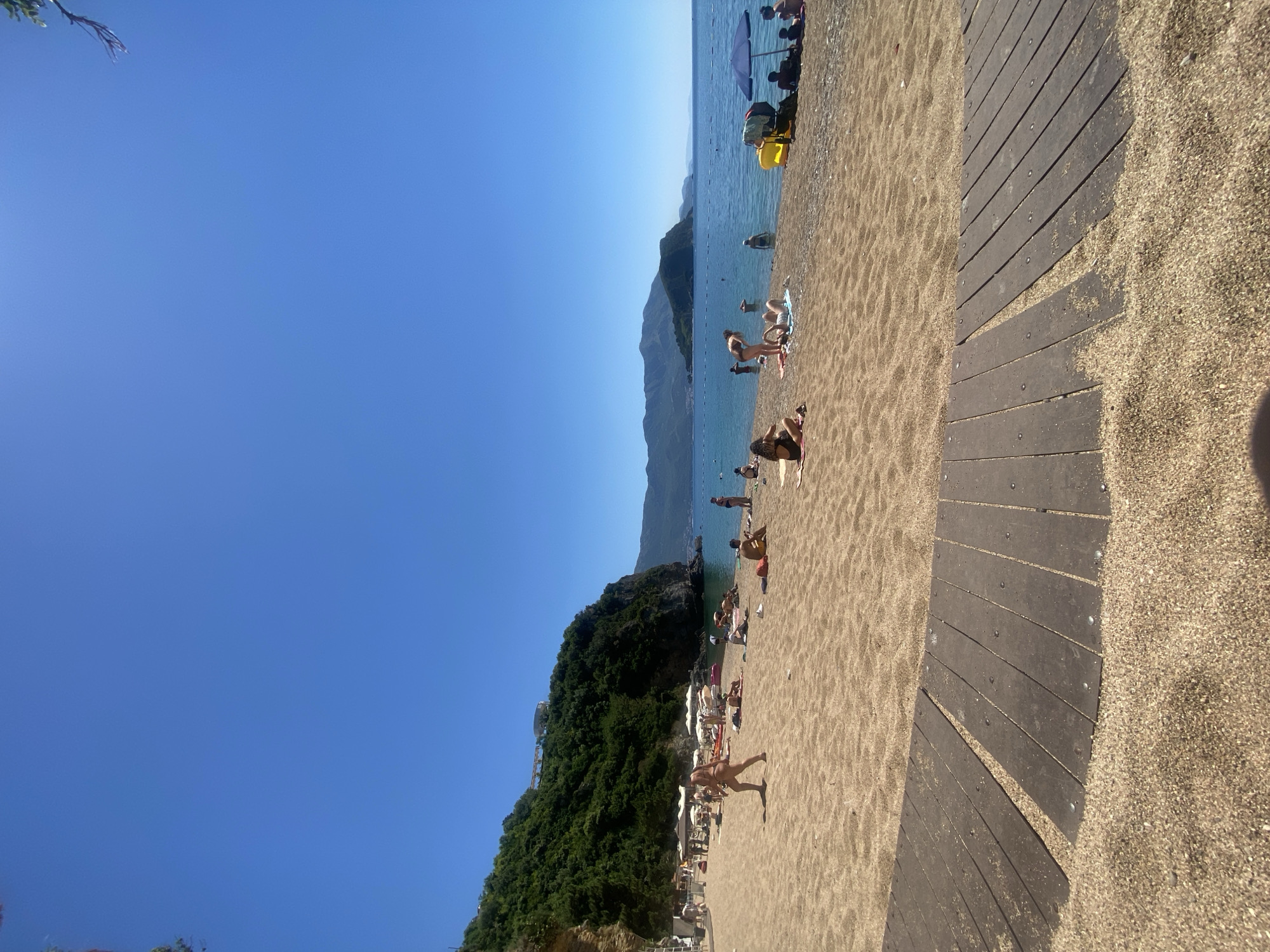 The image shows a beach in Budva, Montenegro, on a sunny day. People are sunbathing and swimming. A wooden walkway leads along the beach. Mountains are visible in the background. Lush green vegetation is on one side of the beach. The sky is clear and blue.