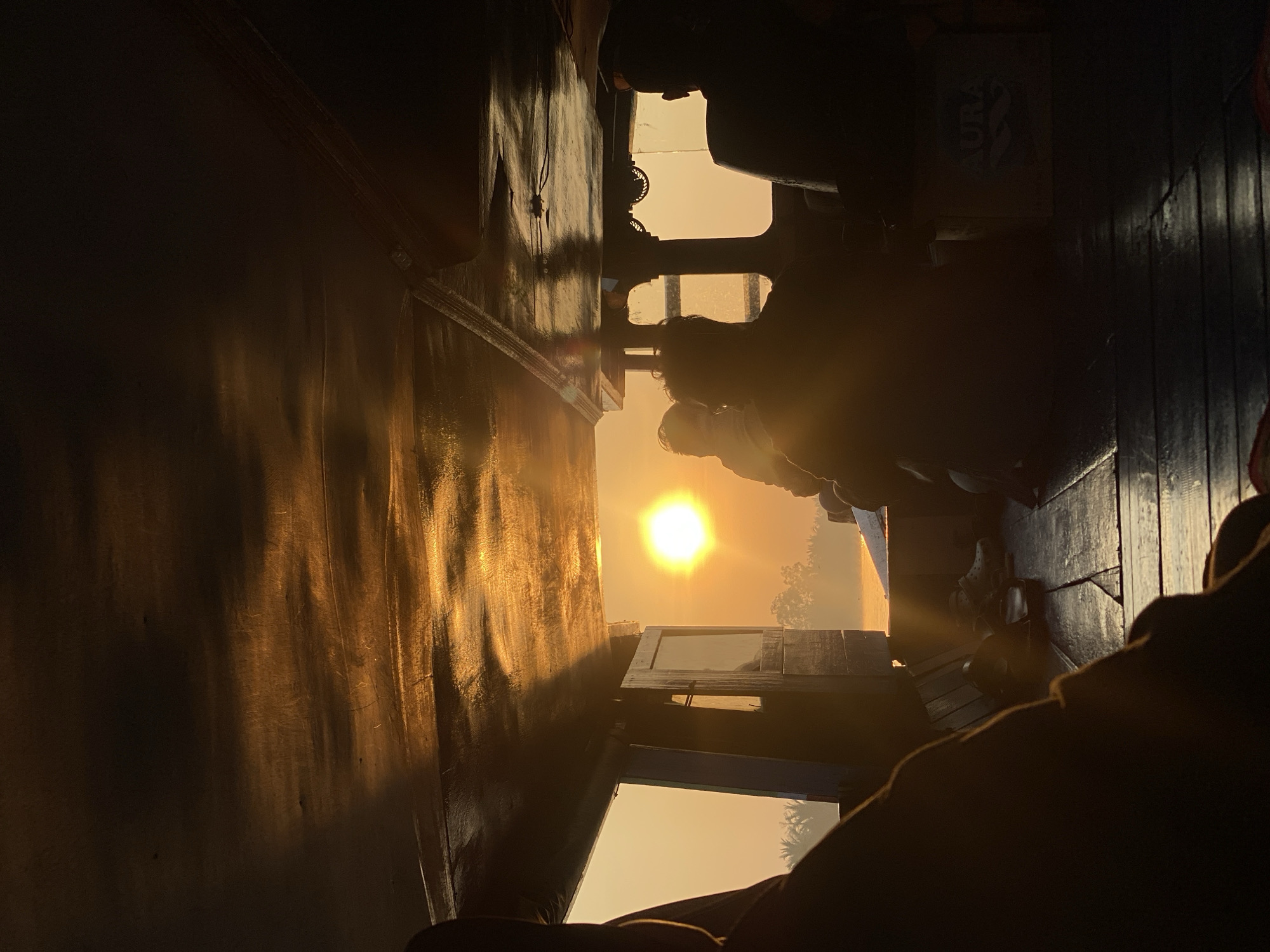 The image depicts a view from inside a wooden boat at sunrise or sunset, possibly on the river in Sungai Tandipah, Indonesia. The sun, a bright orb, is visible in the distance, casting a warm glow over the water and the surrounding foliage. Several individuals are seated inside the boat, silhouetted against the light. The interior of the boat features a wooden ceiling and floor. In the foreground, a person's leg is visible, suggesting the photographer is also on board. The overall atmosphere is tranquil, with the natural light creating a serene and picturesque scene.