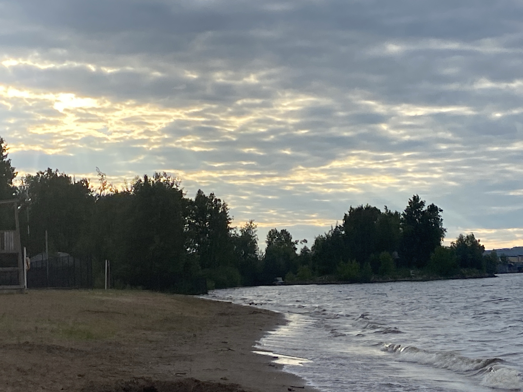 The image depicts a tranquil scene along the shoreline of the Kama River in Chaikovsky, Russia. The foreground features a sandy beach with some scattered debris. The water is gently lapping at the shore, with small waves visible. Beyond the water, a dense line of trees forms the backdrop, with a few structures and buildings partially visible through the foliage on the far right. The sky is overcast with dramatic clouds, illuminated by the golden hues of a sunset or sunrise peeking through breaks in the clouds. The overall atmosphere is serene and natural, characteristic of a riverside landscape.