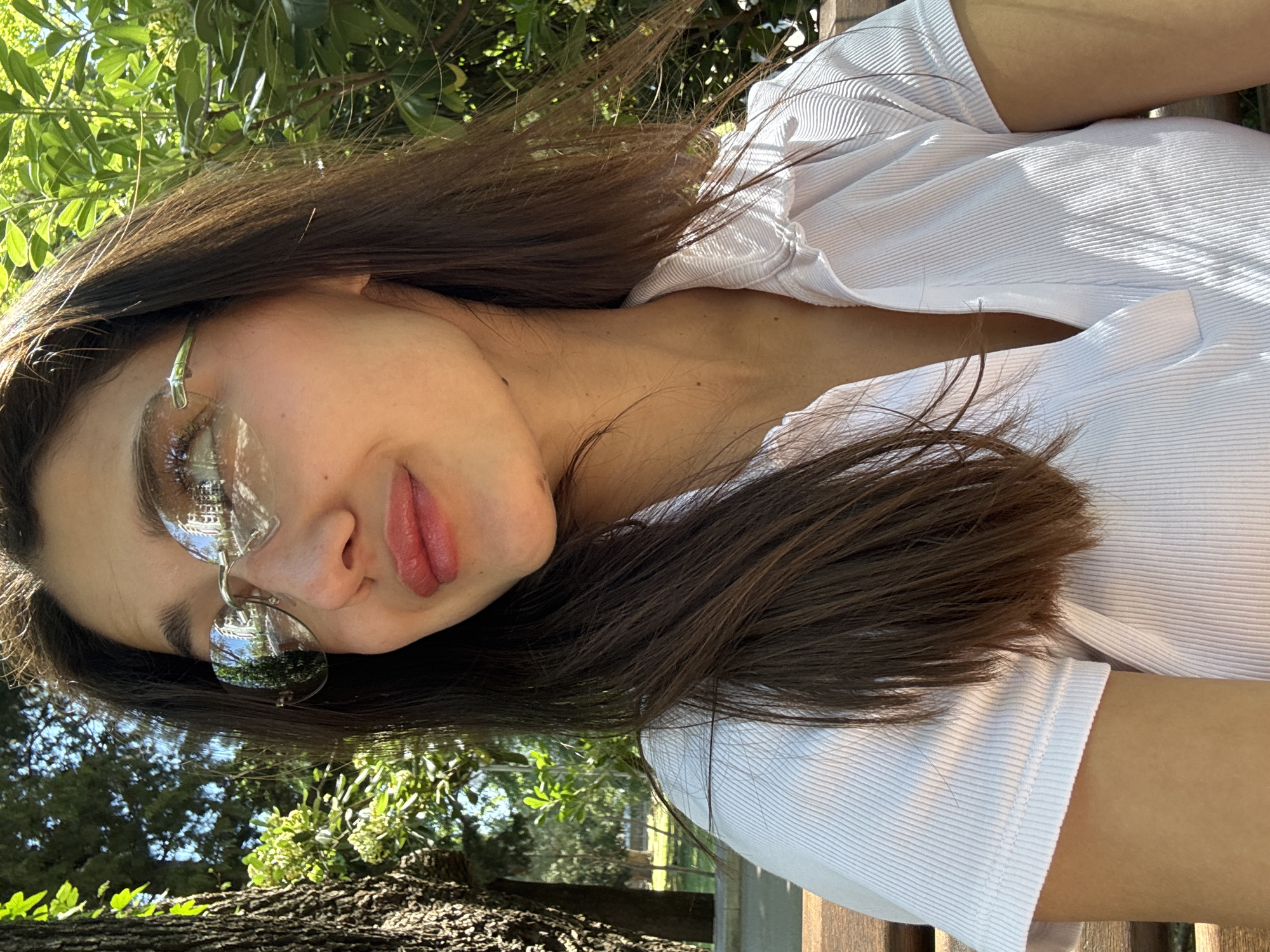 A woman with long brown hair is positioned in an outdoor setting, possibly a park, suggested by the presence of trees and greenery in the background. She is wearing a white, ribbed shirt and reflective glasses. The location appears to be in Bar, Montenegro, based on visual cues. The setting includes natural elements like trees and grass, indicating a park or garden-like environment. The woman is the central subject, and the setting provides context to the scene.