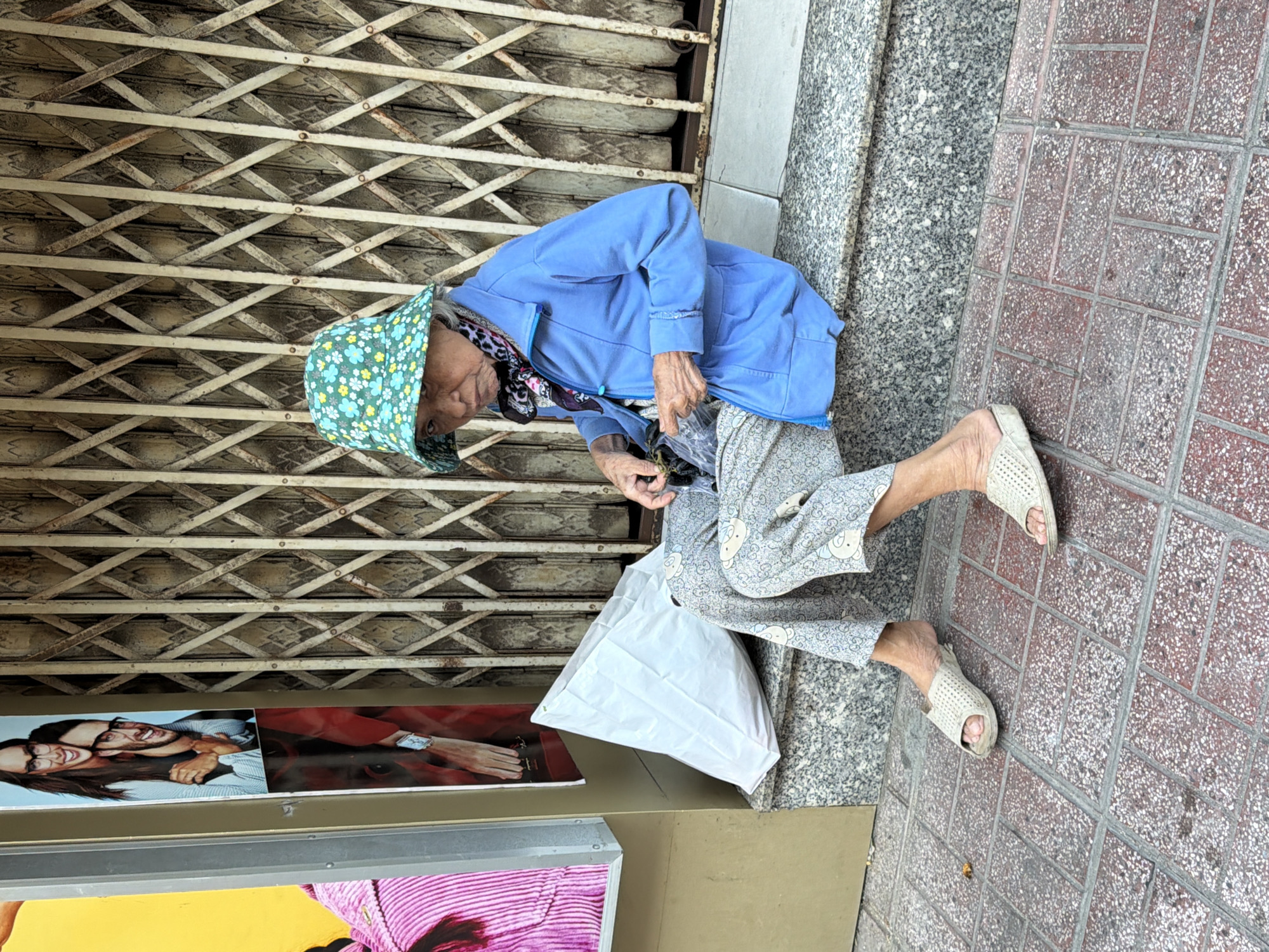 An elderly woman wearing a blue jacket, a floral bucket hat, and patterned pants sits on a granite step in front of a metal security gate. She holds a dark object in her right hand and appears to be examining it. To her left, a large white plastic bag rests on the step. In the background, a poster displays images of people wearing glasses. The scene is located on a sidewalk in Nha Trang, Vietnam.