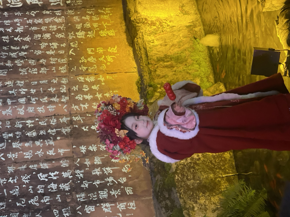 The image displays a young child, appearing as a girl, positioned centrally, facing slightly right of the viewer. She is dressed in a traditional-style deep red robe with white fur trim and an elaborate headpiece adorned with red, orange, and yellow artificial flowers. Her right hand grasps a red, multi-sectioned object, consistent with a candied fruit skewer. The background consists of a rugged rock wall, illuminated from below by warm yellow light, featuring extensive white Chinese calligraphy inscribed in vertical columns. In the lower mid-ground, a pond or shallow pool reflects the yellow light, containing several small orange fish and a narrow fountain jet. Green foliage is visible in the lower left foreground near the water. A black stage lighting fixture rests at the pond's edge. The scene is located in Yuzhong District, China.