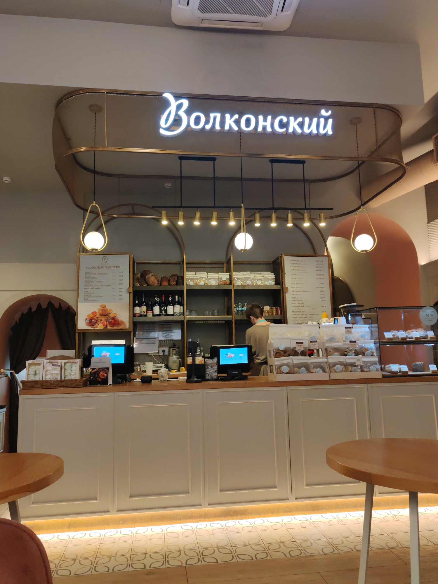The image presents the interior of a "Волконский" (Volkonsky) cafe or bakery, a bright and modern space, likely in Saint Petersburg, Russia, as indicated by the visible text. The central focus is a long service counter behind which a staff member is visible, performing tasks.
Above the counter, a prominent neon sign spells "Волконский" in Cyrillic. The area is well-lit by stylish pendant lights with spherical bulbs and a track lighting system, all featuring gold-colored accents. The counter itself is white with paneled detailing and a light wood top, holding various baked goods in clear display cases. One display case shows a " -50%" discount sticker.
To the left, a large menu board lists "СЕЗОННЫЕ НАПИТКИ" (Seasonal Drinks), including items like "БРУСНИКА", "КЛЮКВА", "ОБЛЕПИХА", "ИМБИРЬ ЛИМОН", "СМОРОДИНА БАЗИЛИК", "ПРЯНЫЙ КОФЕ", and "КАКАО", each with prices in rubles. Another menu board on the right details coffee and tea options such as "ЭСПРЕССО", "ЛАТТЕ", "КАПУЧИНО", "ВОДА", "ТРАВЯНОЙ ЧАЙ", and "ФРУКТОВЫЙ ЧАЙ". Shelving behind the counter displays bottles, possibly wine or juices, and packaged goods.
In the foreground, two round wooden tables with light-colored legs suggest a customer seating area. The floor features a decorative tiled pattern with repeating organic shapes in light tones, complemented by a warm, indirect lighting strip along the base of the counter. Architectural elements include a rounded archway on the left and a curved, peach-colored wall section on the right, contributing to a contemporary and inviting ambiance. The scene depicts a functional, clean, and aesthetically pleasing cafe environment.