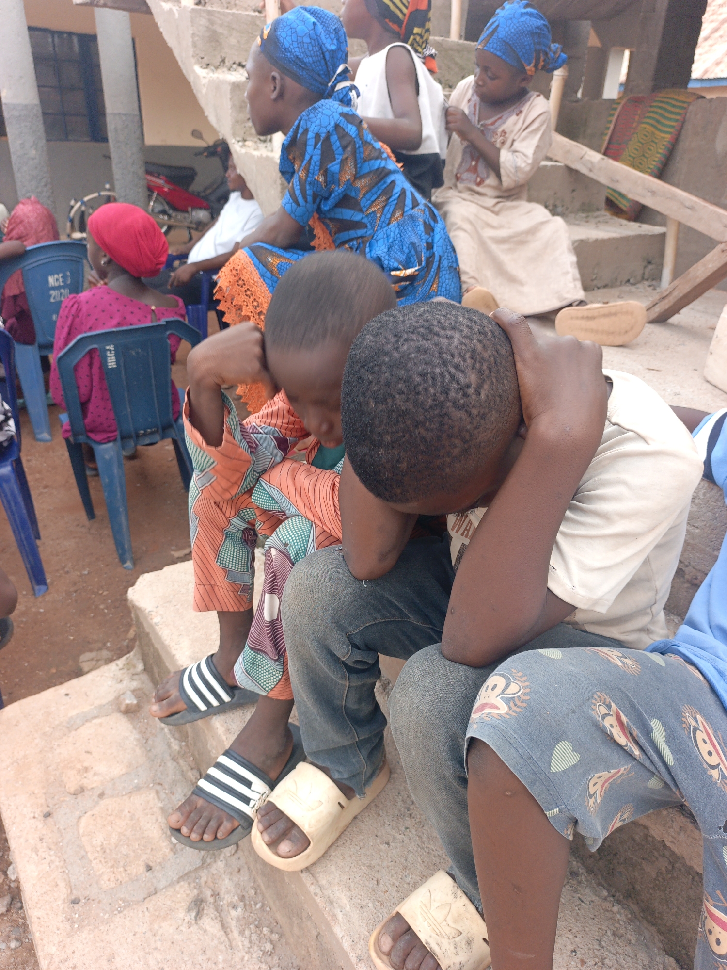 The image captures a gathering of people, primarily children, in Akwanga, Nigeria. In the immediate foreground, two boys are seated on concrete steps. One boy on the left, wearing patterned orange and green clothing with striped sandals, has his head down with his hands near his face. Beside him, another boy, wearing a light shirt and jeans with cream-colored slides, has his head in his hands, resting on his knee, suggesting distress or exhaustion.
Behind them, several other children and adults are visible. A girl in a blue patterned headwrap and matching dress sits on the concrete steps. To her left, another girl in a bright pink top and red headwrap is seated on a blue plastic chair. Other children in traditional attire and headwraps are scattered on the steps and chairs. An adult, partially visible, also sits on a blue plastic chair further back.
The setting appears to be an outdoor or semi-outdoor communal area with concrete steps and dirt ground. In the background, part of a building is visible, along with a red motorcycle/moped parked. A colorful patterned mat or fabric is draped over the steps in the midground. The lighting suggests it is daytime, likely sunny.
Visible text includes "NCE 2020" and "HOCA" printed on the backs of the blue plastic chairs, and an Adidas logo on the slides worn by the boy on the right. The overall atmosphere suggests a communal gathering or event, with some participants showing signs of weariness.