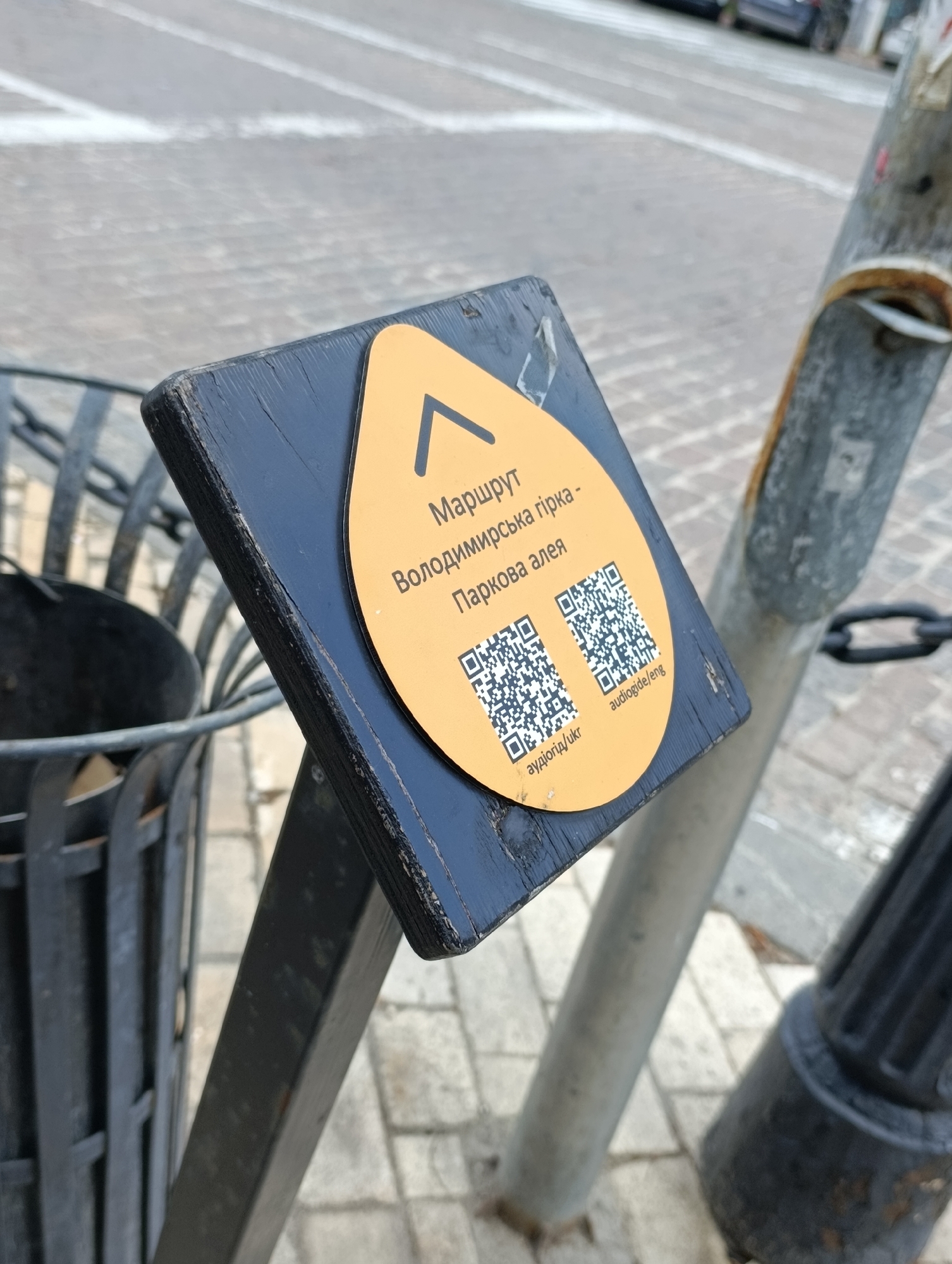 A close-up shot shows a sign with a yellow, oval-shaped sticker attached to a black, wooden post. The sticker has a black arrow pointing upwards in the top left corner. Below the arrow are lines of text in Ukrainian, reading "Маршрут" (Route), "Володимирська гірка" (Volodymyrska Hill), and "Паркова алея" (Park Alley). Two QR codes are printed on the sticker: one labeled "аудіогід/ukr" (audio guide/ukr) and another labeled "audiogide/eng" (audio guide/eng). The sign is positioned next to a black metal trash can with a lattice design. In the background, a paved street and a metal pole with a chain are visible, suggesting an outdoor urban setting, likely in Kyiv, Ukraine.