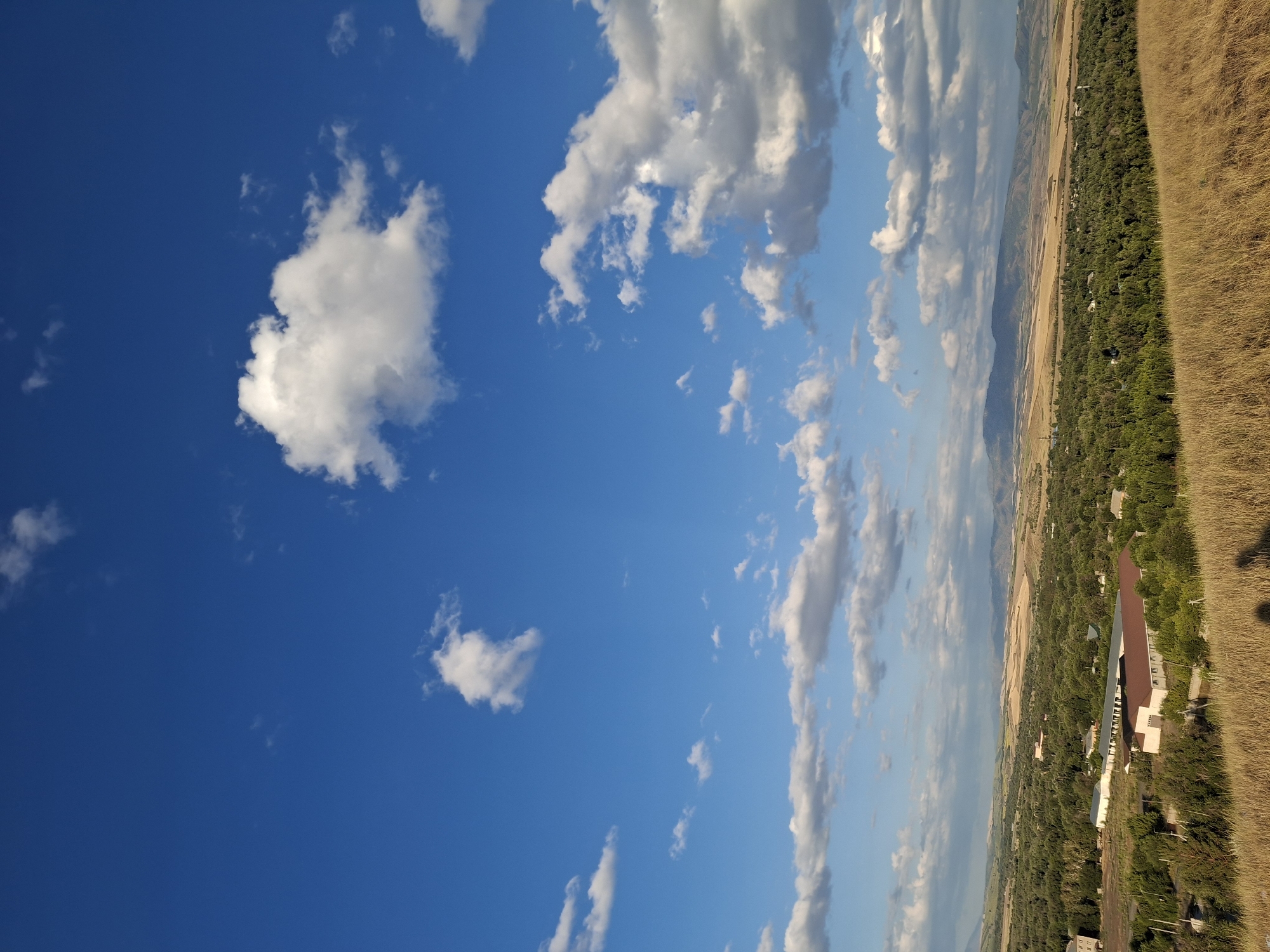 The image depicts a rural landscape with a bright blue sky dotted with white clouds. In the foreground, dry, golden grass covers a hillside. Below the hill, a treeline of lush green trees separates the foreground from a cluster of buildings. One prominent building has a reddish-brown roof and white walls. Further in the distance, rolling hills and fields of a lighter, tan color stretch towards a mountain range under the expansive sky. The overall scene suggests a sunny day in a natural setting, possibly near a rural settlement. There is no visible indication of Kabanbay, Kazakhstan.