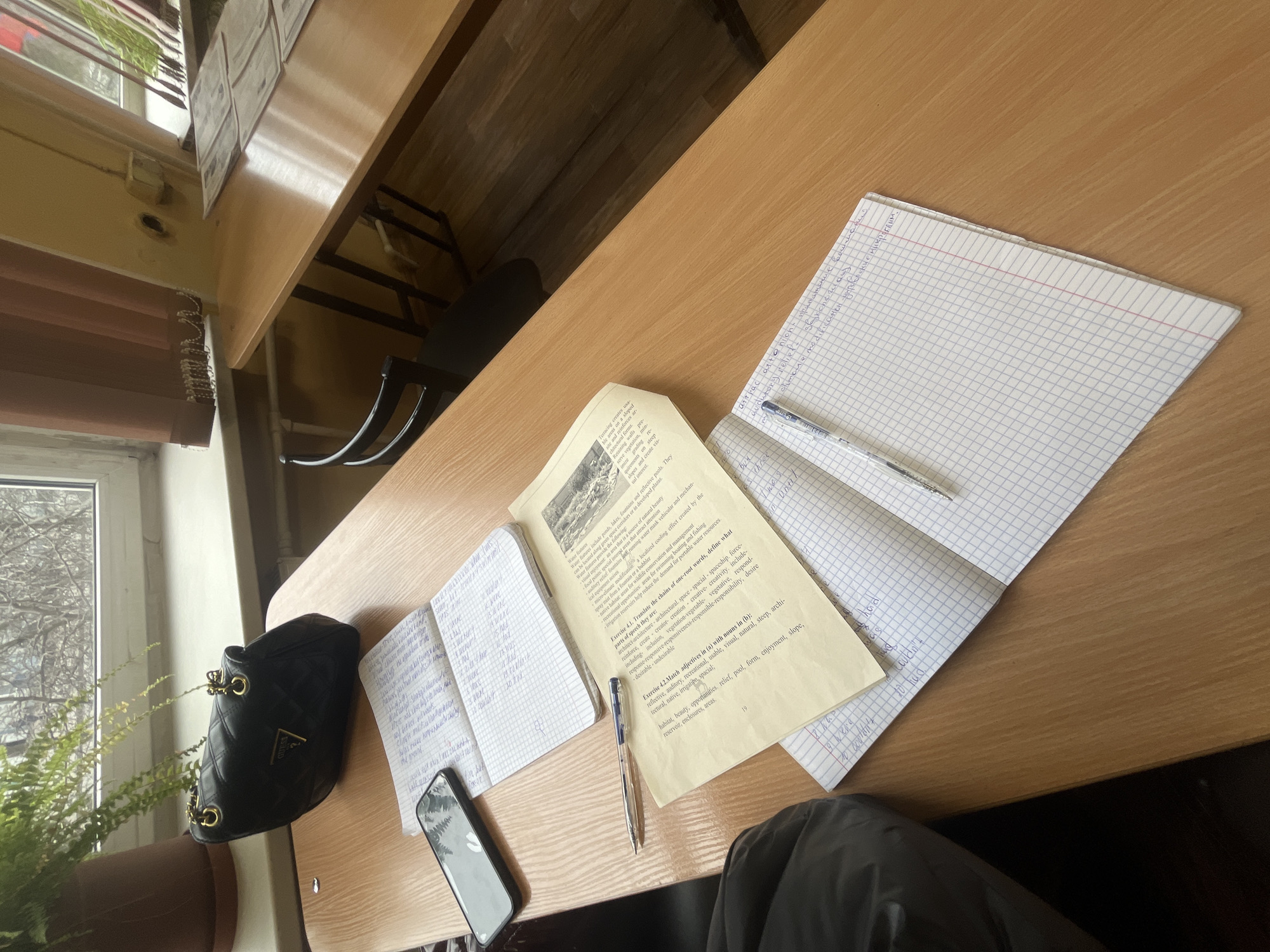 The image shows a desk or table in a classroom, likely in Saint Petersburg, Russia, judging by the appearance of the windows and surrounding structures outside.
On the desk, there are several open notebooks, one containing printed text and the other hand-written notes in the Russian language. A blue pen lies on top of one of the notebooks. A black Guess purse and a smartphone are also placed on the desk. A person wearing dark clothing is seated at the desk, near the bottom right of the frame. A potted plant is visible by the window. The desk appears to be made of light-colored wood. The scene suggests a student studying or working on assignments.