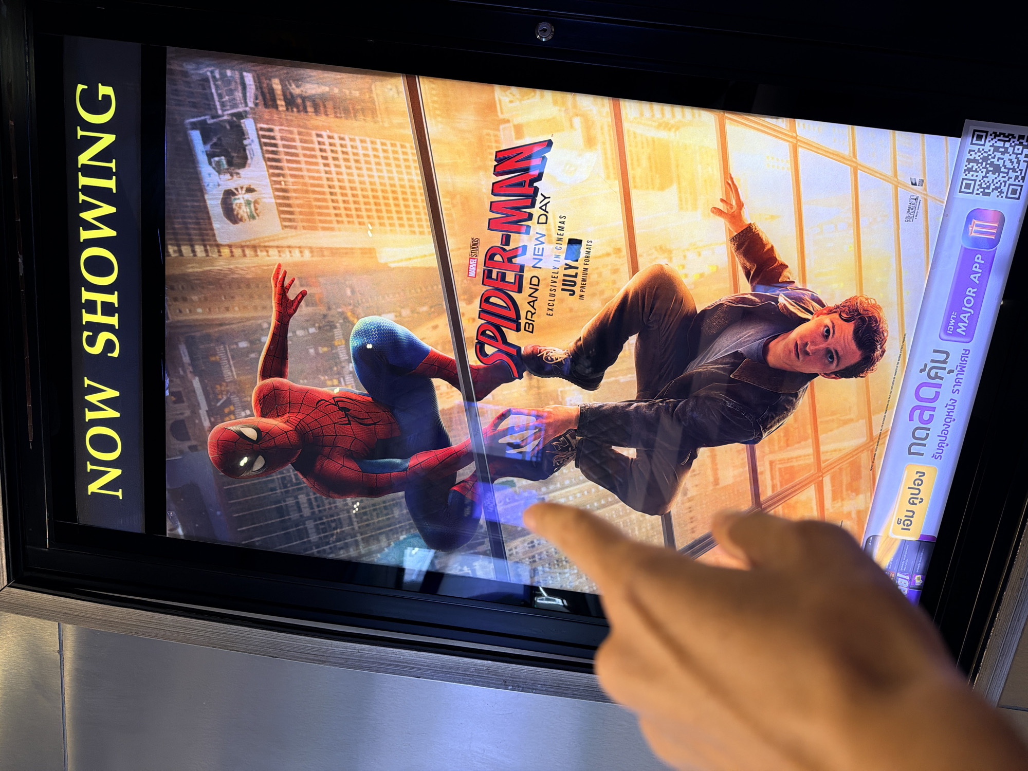 A person's finger points at a movie poster displaying "SPIDER-MAN BRAND NEW DAY" and "JULY" in a cinema lobby. The poster features Spider-Man in his costume and an upside-down reflection of Peter Parker. The text at the bottom of the poster is in Thai and reads "Get discounted movie tickets" and "Exclusive to Major App." A QR code is visible next to the text. The scene suggests a movie theater environment, likely in the city of Ko Samui District, Thailand, given the Thai text.