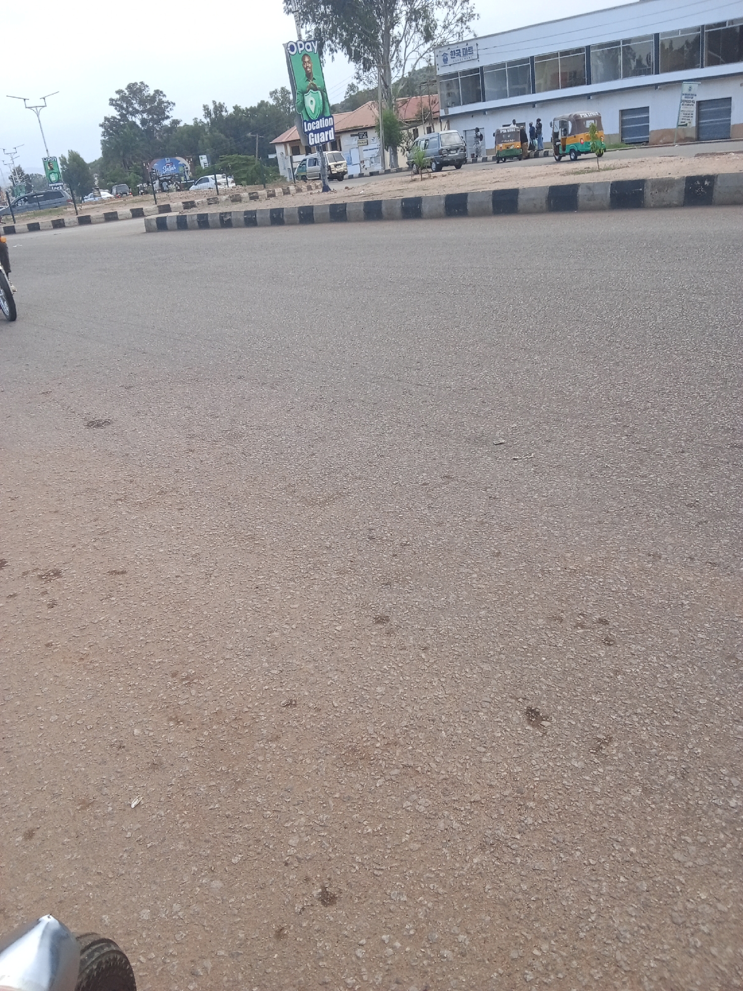 The image shows a street scene in Gura Top, Nigeria. In the foreground, the asphalt road takes up most of the frame. In the midground, there is a raised median with black and white striped borders. Beyond the median, several vehicles are visible, including a gray van, two auto-rickshaws (one yellow and green, the other brown and green), and several parked cars. People are standing near the vehicles and on the sidewalk. A white building with large windows and signs, including one with Korean script and "화 한국 마트," is in the background. There are also trees and utility poles along the street. A large banner featuring a man and the text "OPay Location Guard" is also visible. The sky is overcast.