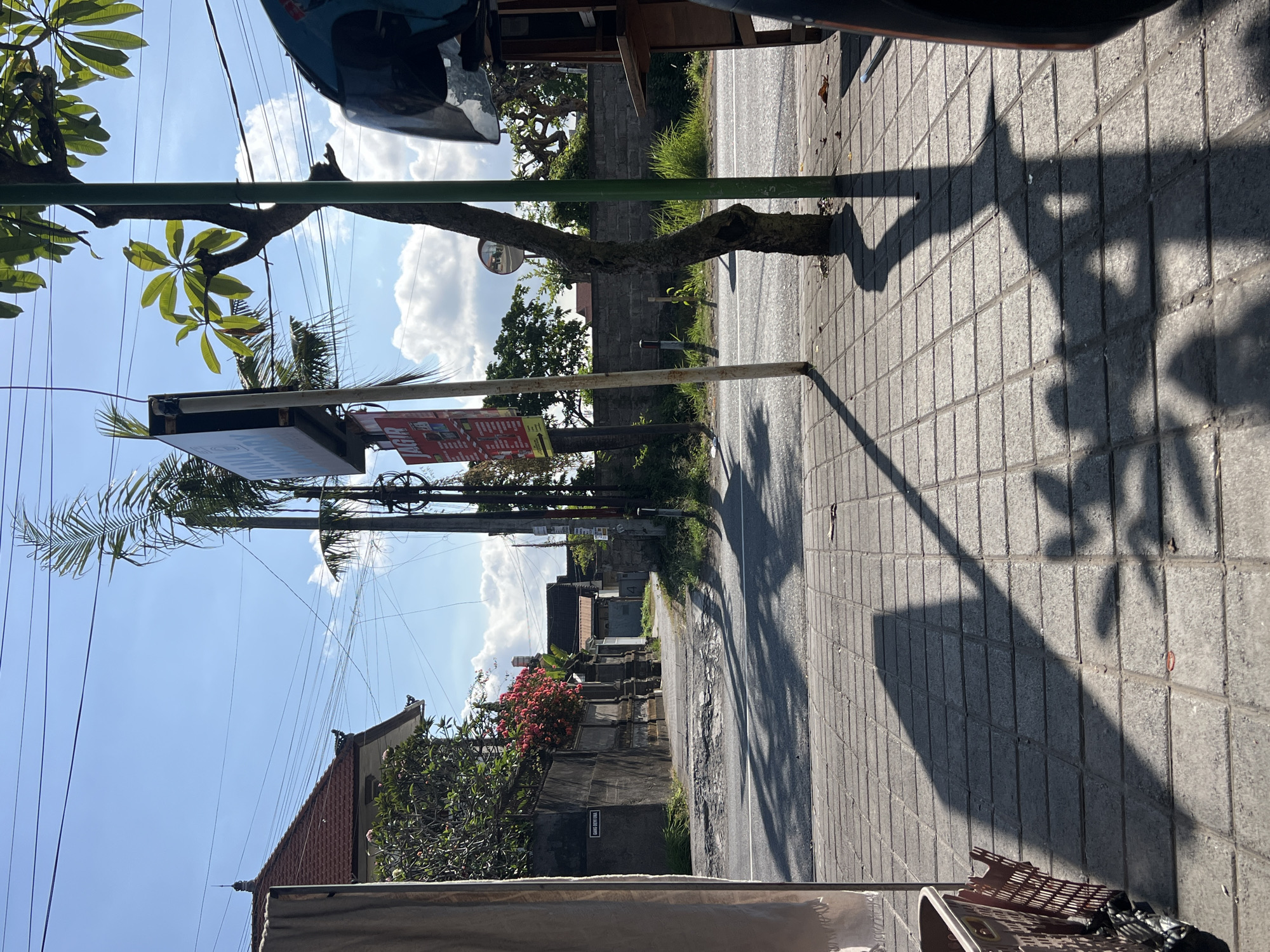 This image captures a street scene in Denpasar, Indonesia, on a clear day with bright sunlight casting strong shadows. The foreground is a tiled sidewalk with a shadow from a tree and possibly a shopping basket on the left. To the right, a portion of a motorcycle is visible, including a blue helmet. A narrow, paved road runs alongside the sidewalk, and in the distance, a street curves to the left, lined with traditional Balinese houses with red tiled roofs and stone walls. Lush green vegetation and flowering bushes, including a vibrant red bush, are present along the walls. Several utility poles with numerous wires crisscross the sky, and a tall palm tree stands near a large signpost. The signpost has a prominent advertising poster on it. The overall impression is of a typical, sunny street in an urban area of Denpasar, likely a residential or mixed-use neighborhood.