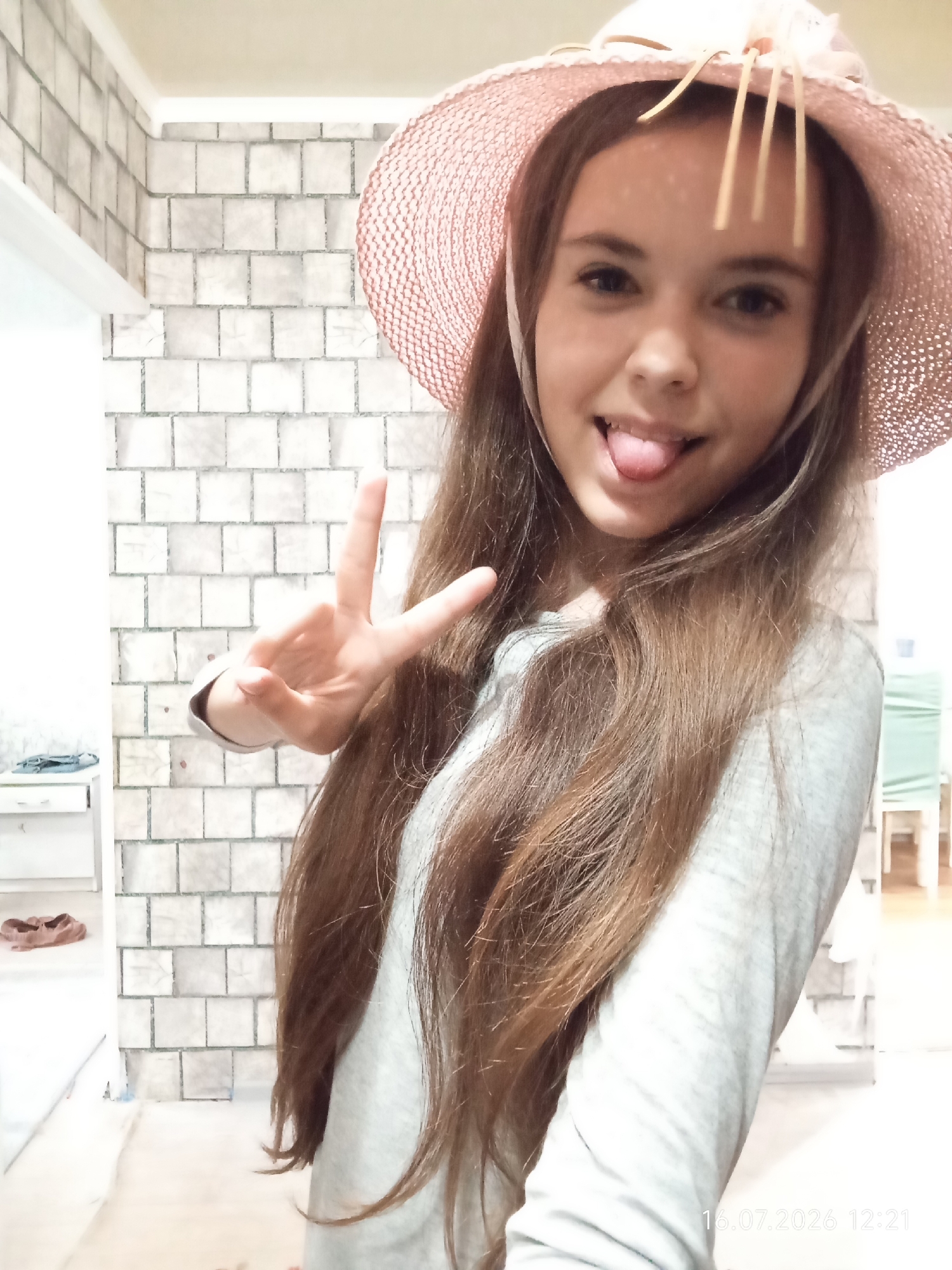 A young female with long brown hair and a pink straw hat is pictured from the chest up, posing for a selfie. She is sticking her tongue out and making a peace sign with her left hand. She is wearing a light gray long-sleeved shirt. The background appears to be an indoor setting with a white tiled wall. There is a white shelf unit with a pair of brown shoes on it to the left of the frame. The image has a timestamp in the lower right corner indicating "16.07.2026 12:21". This image was taken in the city of Qobda, Kazakhstan.