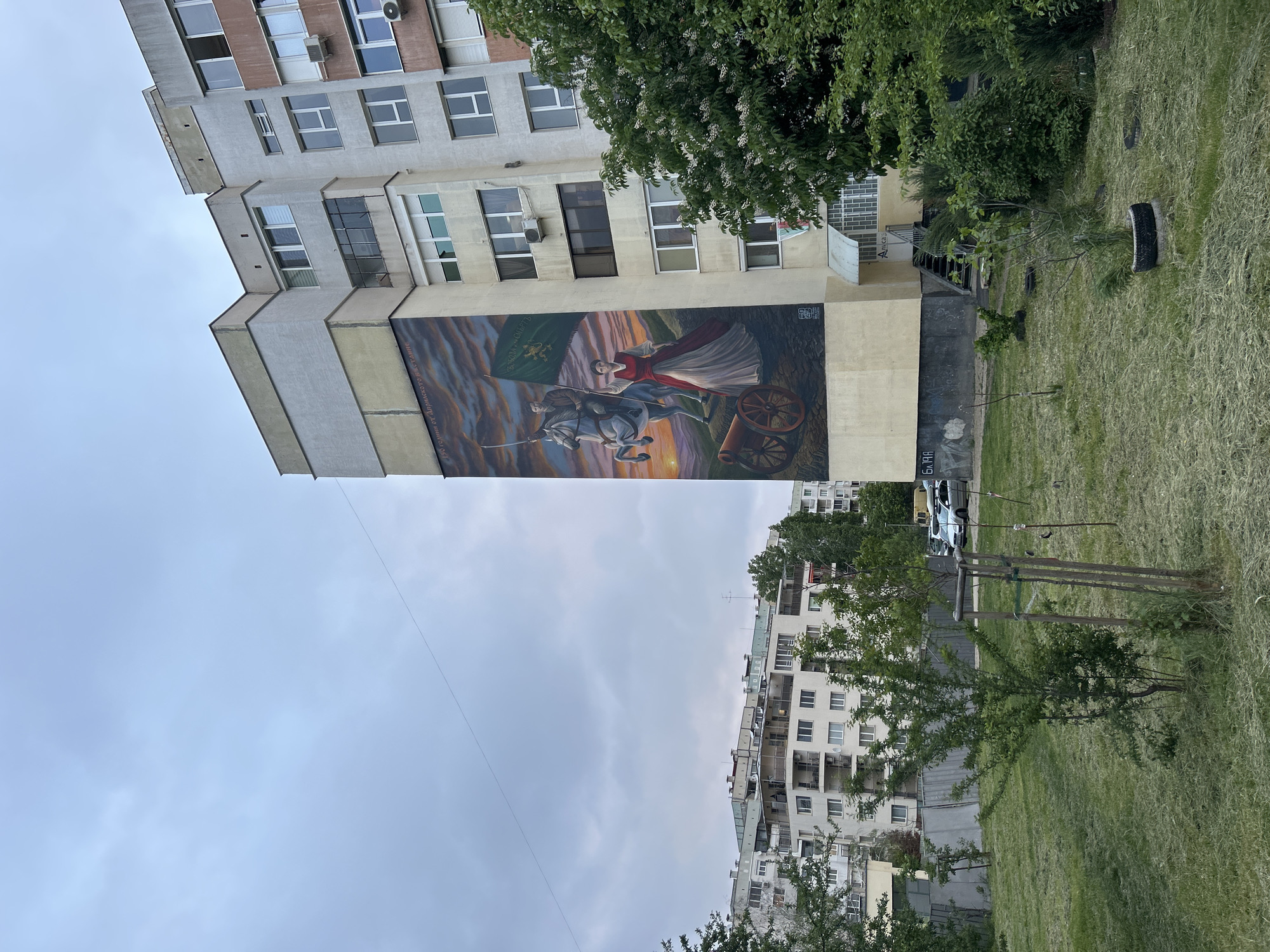 The image shows a mural painted on the side of a building. The mural depicts a man on horseback holding a flag, a woman in a red dress, and a cannon. The sky in the background is orange and yellow, suggesting a sunset or sunrise. The building is light grey and has several windows. In front of the building is a grassy area with trees and used tires. In the background, there are other buildings and a cloudy sky. The location appears to be in an urban area.