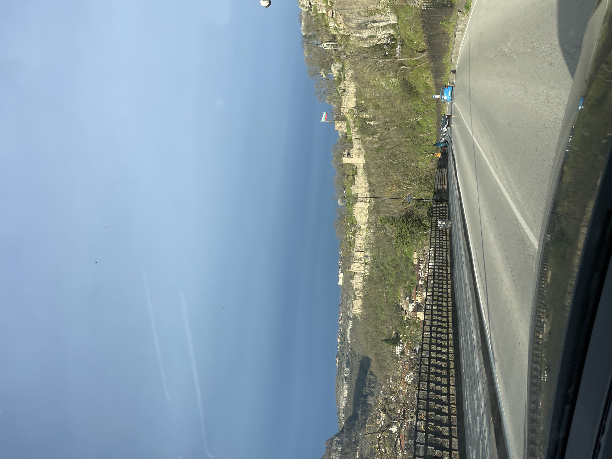 The image shows a view from a vehicle traveling on a road alongside a cliff in Veliko Tarnovo, Bulgaria. The road is gray asphalt with a white dividing line. A black metal fence separates the road from the cliff edge. Below the cliff is a cityscape of houses and buildings. At the top of the cliff are stone fortifications and vegetation. A Bulgarian flag is visible on the fortifications. There are a few vehicles and people on the other side of the road. The sky is blue with light clouds.