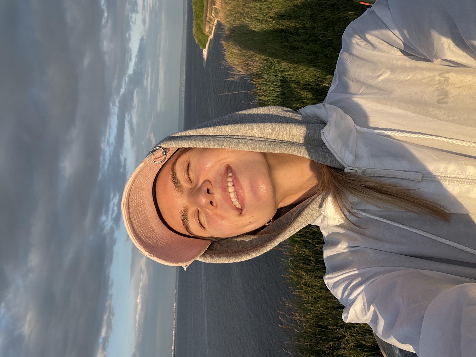 A young woman is captured in a sun-drenched selfie outdoors, positioned on a grassy cliff overlooking a vast body of water. She wears a light pink baseball cap backwards, a gray hooded sweatshirt, and a translucent white windbreaker jacket with a faint "NASA" logo on the lower right side (viewer's left) and a drawstring visible. Her eyes are closed, and she displays a broad smile, with her head tilted slightly upwards and to her right, enjoying the warm light. Small gold hoop earrings are visible on both ears, and a silver-toned metal ring adorns the brim of her cap.
The background features a large expanse of dark blue water, which could be a river or lake, stretching horizontally across the mid-ground. On the distant horizon across the water, a faint skyline with what appears to be urban structures is visible. To the right, a sandy or light-colored cliff face meets the water. The sky above is partially covered with dark gray clouds, interspersed with patches of lighter blue, all illuminated by the warm, golden light of either sunrise or sunset, casting long shadows from the foreground grass. The immediate foreground consists of dry, tall grasses and green vegetation on the cliff edge.