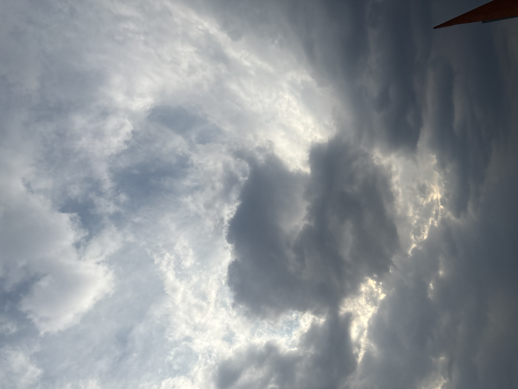 The image shows a cloudy sky, with a mix of dark gray and white clouds. Some clouds are dense and block the sunlight, while others are thin and allow light to filter through. The sky is shot from a low angle. An orange object is visible in the top right corner of the frame. This photo was taken in Mexico City, Mexico.
