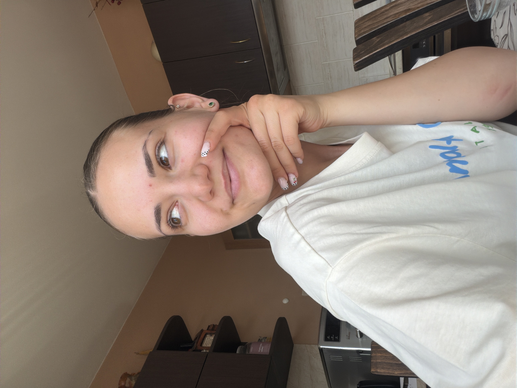 A young woman is depicted indoors, likely in a kitchen or a room with a kitchen area, indicated by the dark brown cabinetry with gold handles, a wooden surface, and a visible metal appliance in the background. The walls are a light brown or beige color. She is looking directly at the camera with a slight, gentle smile. Her dark hair is pulled back, and her left ear has two visible piercings: a small diamond-like stud in the lobe and a green stud in the helix. Her fingernails are manicured, featuring white tips with black polka dots on some, and small floral designs on others. She wears a light-colored, possibly off-white, t-shirt with partially visible blue text that includes "ITAL" and other unclear letters. Her left hand is casually resting on her cheek, with an index finger near her mouth. The lighting suggests it is daytime, and the overall mood is calm and relaxed.