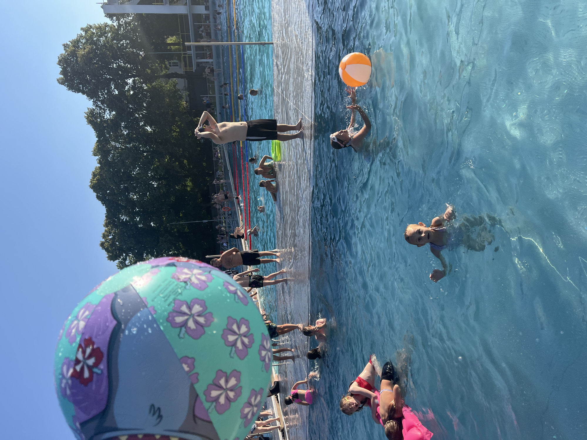The image depicts a crowded outdoor swimming pool on a sunny day. In the foreground, a large inflatable ball with a floral pattern partially obscures the view. The pool is filled with people of various ages swimming and wading. In the middle ground, several individuals are standing on a wet, dark-colored platform that extends into the pool. One man on the platform is wearing swim trunks and has his hands on his head. A young girl in the water is reaching for an orange and white beach ball. In the background, more people are visible in the pool and around its edges. Trees line the perimeter of the pool area, and a clear blue sky is visible above. The overall atmosphere suggests a recreational scene at a public pool.