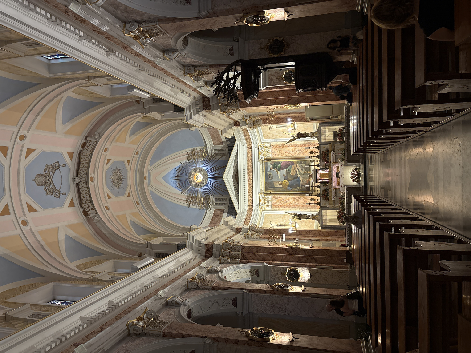Here is a description of the image:
The image shows the interior of St. Peter's Church in Jaffa, Tel Aviv, Israel. The perspective is from within the church, looking towards the altar and the ornate ceiling. The ceiling is a complex design with a central dome featuring a radiating light design and painted with pastel colors. The walls are decorated with marble and intricate carvings. Pews line the sides of the church, and some people are seated in them. The overall lighting is dim, creating a solemn atmosphere.