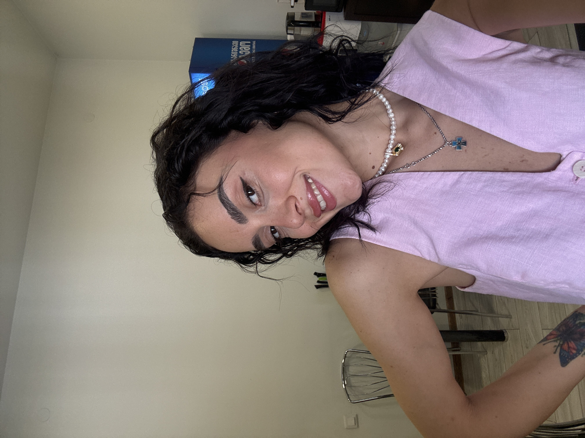 A young woman with dark curly hair and light skin, wearing a pink sleeveless top and a pearl necklace with a cross pendant, poses for a selfie indoors. Her head is slightly tilted, and she is smiling with her eyes looking towards the camera. The background is an indoor setting with a plain wall, a metal chair, and a blue cabinet with text in Georgian script. The lighting appears to be natural, coming from the front. The scene does not provide any specific clues to pinpoint a location within Tbilisi, Georgia.
