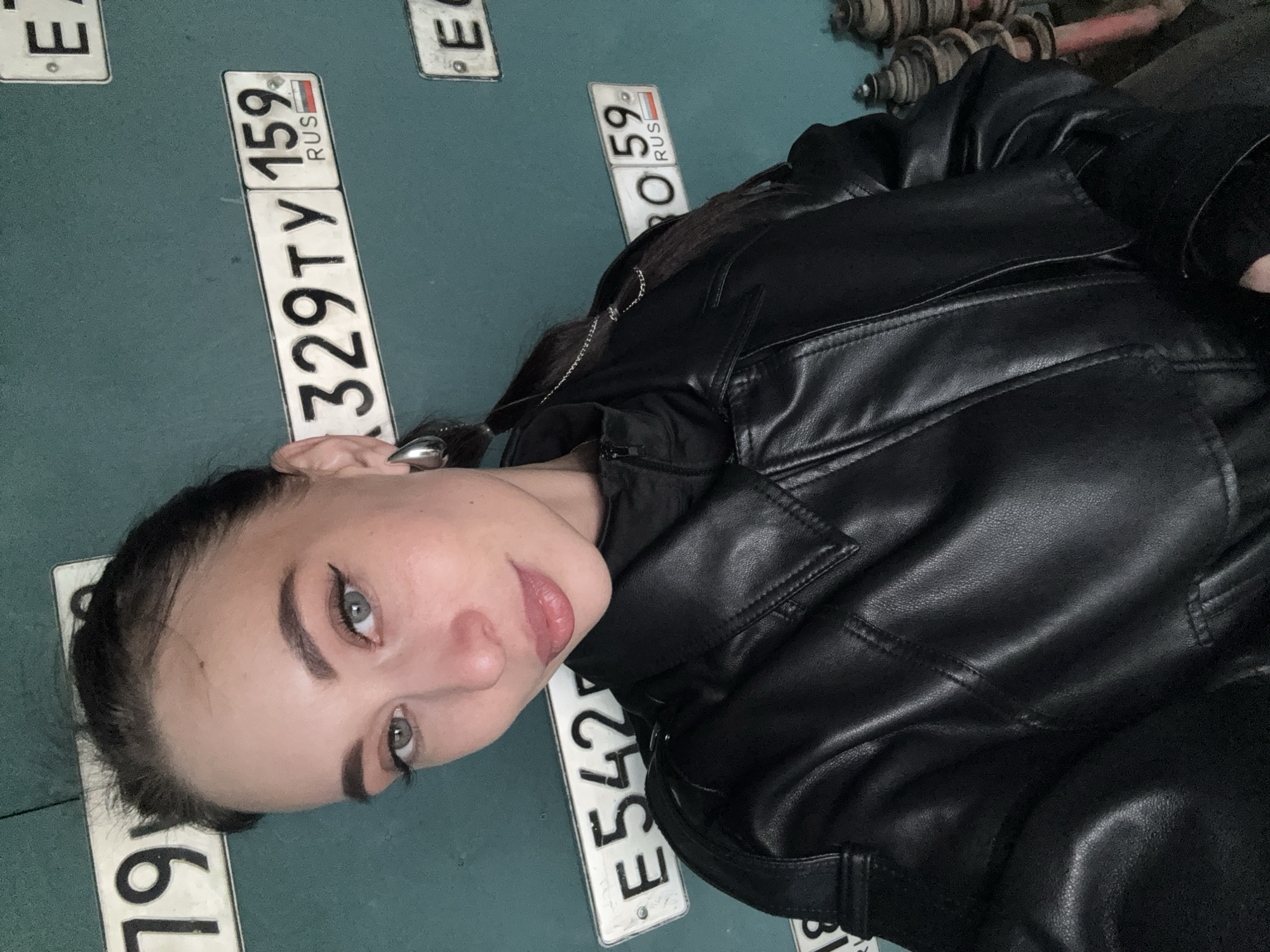 A young woman is captured in a medium-close shot, looking directly at the viewer with a calm and slightly confident expression. Her head is tilted, and her light-colored eyes (likely blue or gray) are prominent, enhanced by winged eyeliner. She has dark hair tied back and well-defined eyebrows. She is wearing a black leather jacket, and a silver-colored earring is visible on her right ear.
The background consists of a teal-colored wall or vertical surface to which multiple Russian license plates are affixed. In the upper right portion of the image, some industrial-looking, possibly rusted, metal machinery or equipment is partially visible, suggesting a workshop or garage setting.
The woman appears to be posing for the photograph. The visible license plates provide clear location cues. The most prominent plate reads "**329 TY 159 RUS**". The "159 RUS" and another partially visible plate showing "**...O 59 RUS**" indicate the Perm Krai region of Russia, which aligns with the provided context of Chaikovsky, Russia. Other partial plate numbers include "**E542...**" and remnants of others like "**E...**" and "**...19...**". The lighting appears artificial, suggesting an indoor environment.