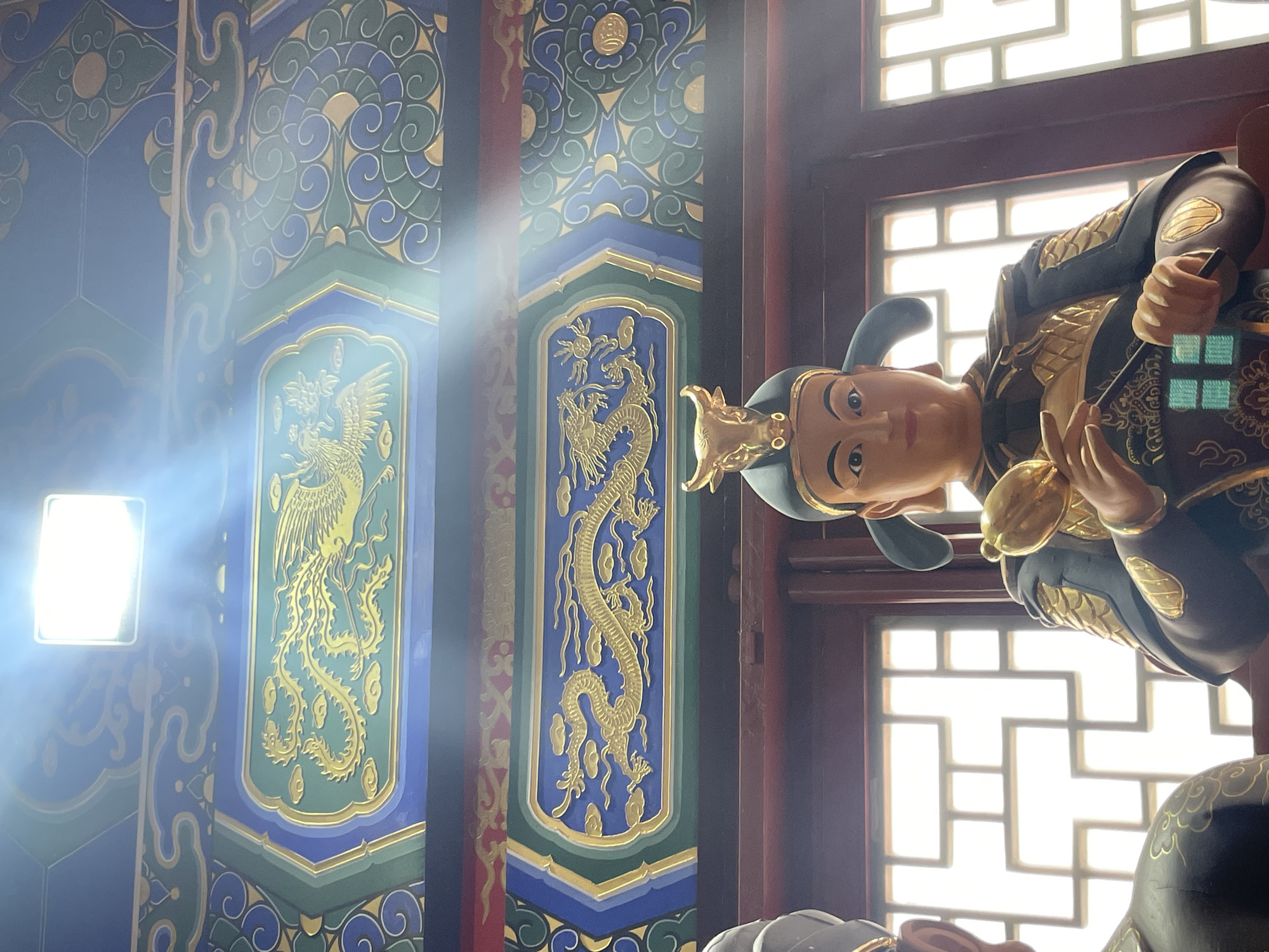 The photograph shows a statue of a person in traditional garb in the city of Lintong District, China. The statue has light skin and is wearing a headdress with golden accents. The statue has on black robes with gold embellishments. The statue is positioned in front of a lattice window with light coming through. Behind the statue, there are ornate columns decorated with phoenix and dragon motifs in gold against a blue and green background. One light is shining brightly and creating a glare.