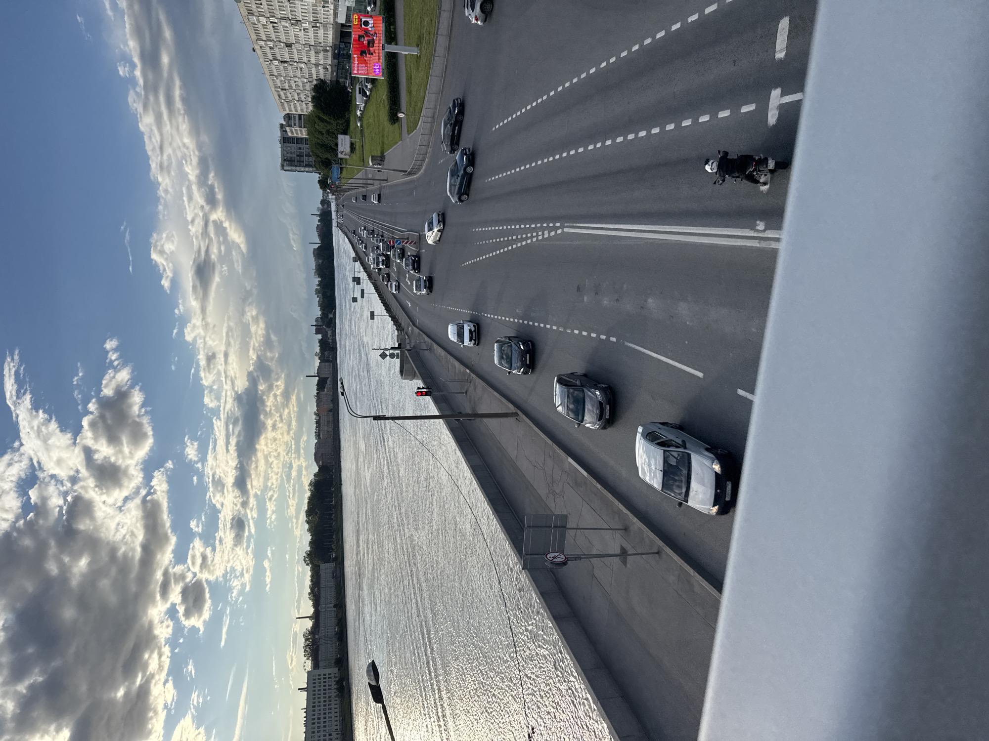This image depicts a view from a bridge over a wide body of water, likely a river or canal, in Saint Petersburg, Russia. The scene is dominated by a multi-lane road filled with traffic, primarily cars and a single motorcycle. The cars are diverse in color and model, with many appearing to be modern sedans. The road itself is well-maintained with clear lane markings, including dashed white lines and solid white lines.
The setting includes urban elements such as tall apartment buildings lining the far bank of the water, with some greenery and smaller structures visible. A large digital billboard with red accents is situated on the left side of the road in the distance. The sky is partly cloudy with dramatic, illuminated clouds, suggesting either early morning or late afternoon. The water is choppy, reflecting the light and creating a shimmering surface.
The overall atmosphere is one of a busy, urban environment during a bright but cloudy day. There is no discernible activity other than the flow of traffic. The perspective is from a high vantage point, looking down and along the road and water. Visible text is limited to what might be on the digital billboard in the distance, which is too small to be legible. The composition emphasizes the linearity of the road and the expanse of the water and sky.