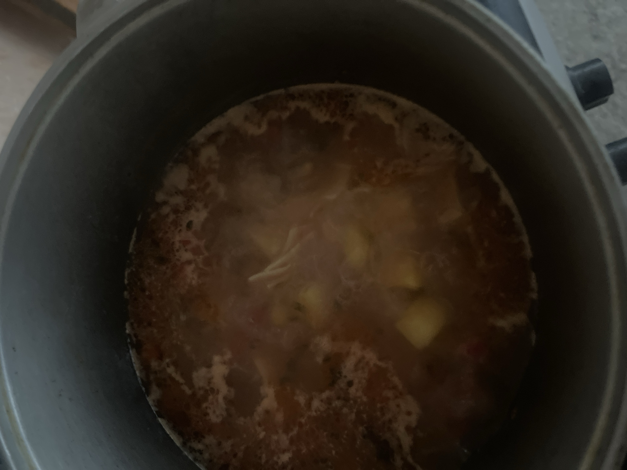 A close-up, overhead shot shows a pot of simmering stew. The stew is a rich, reddish-brown color, with visible chunks of vegetables and what appears to be shredded meat. A light froth sits on the surface, indicating it's actively cooking. The pot itself is metallic and appears to be made of aluminum or stainless steel, showing signs of use. The background is dark and out of focus, suggesting the photo was taken in a kitchen environment. There are no people visible in the image. The scene is indoors.