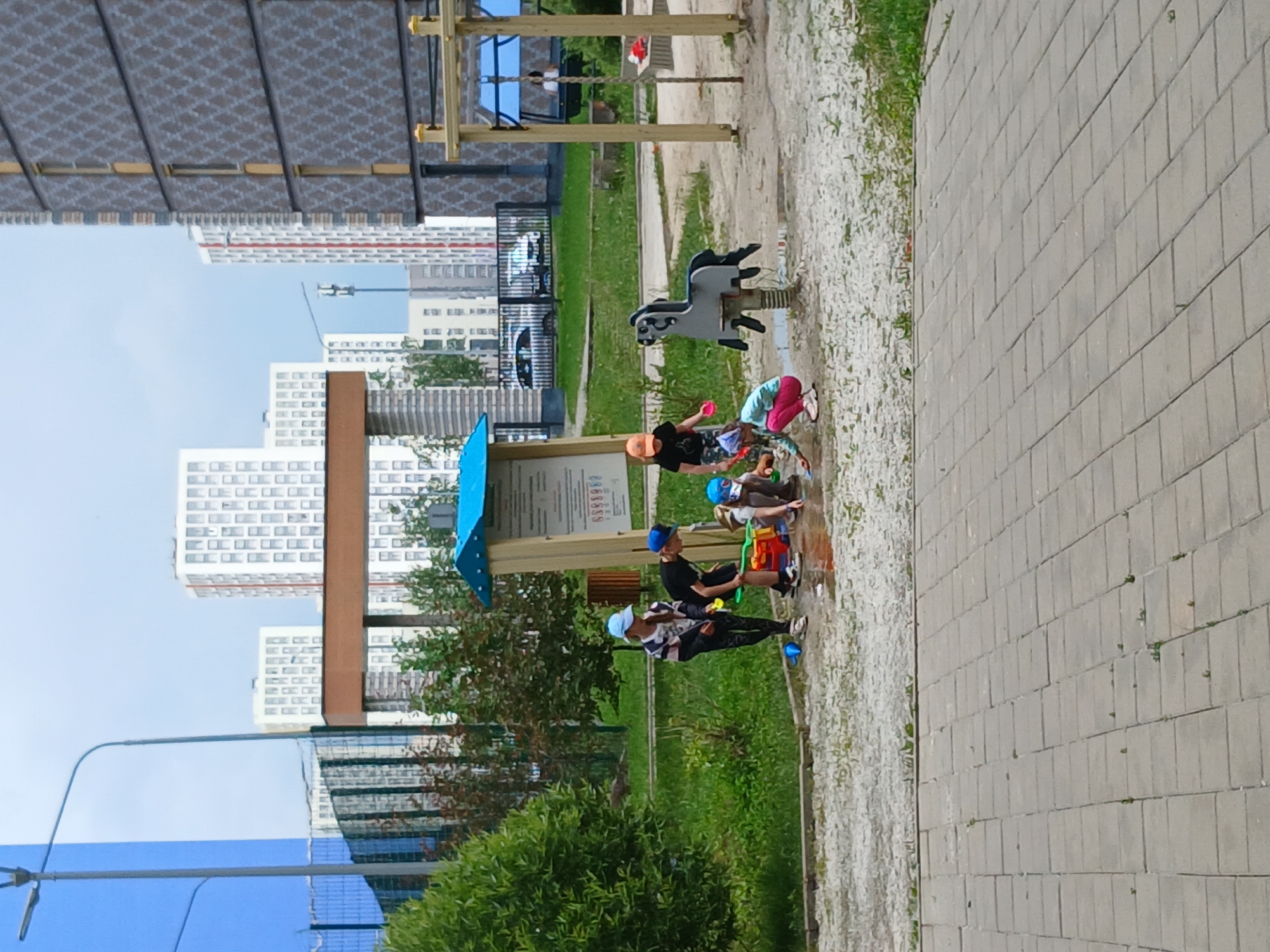 This image depicts a group of children playing in a shallow puddle on a playground in Yekaterinburg, Russia. In the foreground, a paved walkway leads to a gravelly area where several children are engaged in water play. One child, wearing a blue cap and striped shirt, stands and watches, while others are crouched down. Toys, including a bucket and what appears to be a sand toy set, are scattered around the puddle. A spring-mounted rocking horse is visible to the right of the children. In the background, modern multi-story apartment buildings dominate the skyline. The sky is overcast.