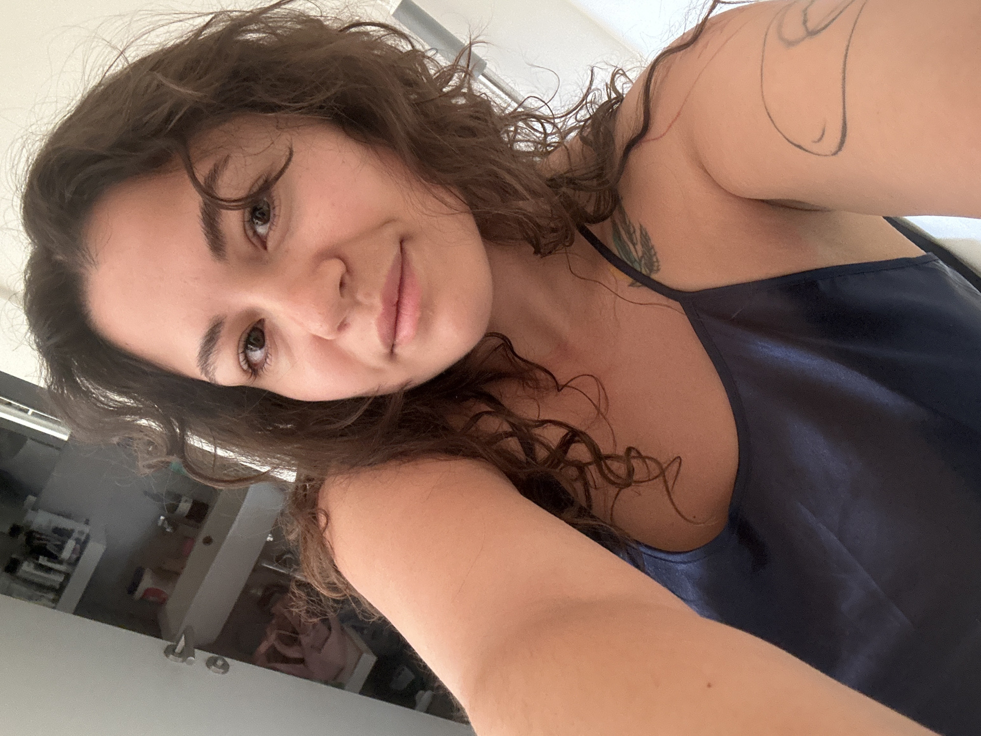 A woman with brown curly hair and a tattoo on her left arm is taking a selfie. She is wearing a navy blue satin top with thin straps. The background appears to be a bathroom or bedroom with light-colored walls. The location in Budva, Montenegro cannot be determined from the image.