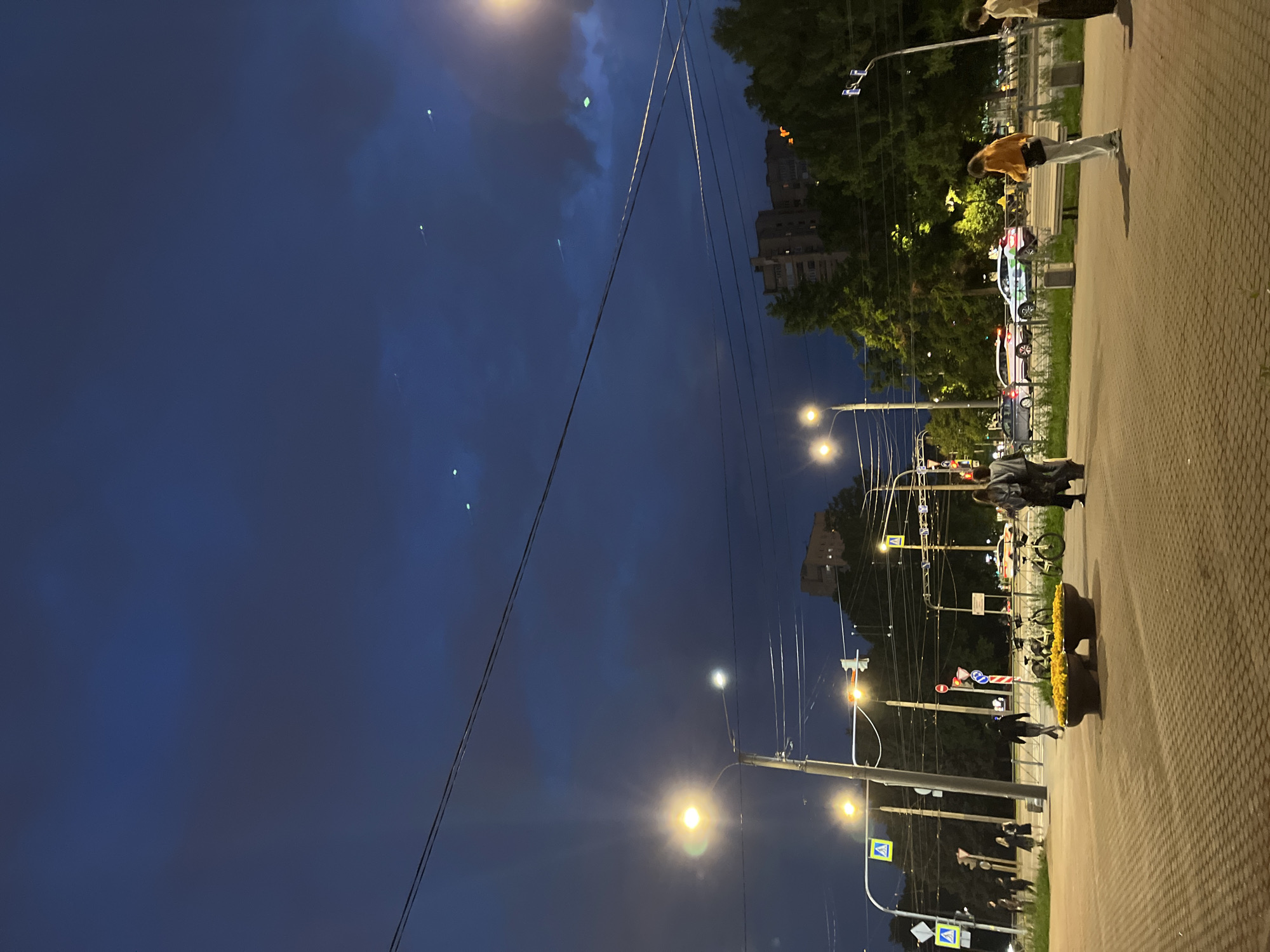 The image depicts a street scene at dusk. Several people are visible walking on a paved sidewalk and crossing the street. One person in the foreground wears an orange jacket and light-colored pants. Another person, wearing a dark hooded sweatshirt and pants, walks across the street. Bicycles are parked near planters filled with yellow flowers. Streetlights illuminate the area, and cars are driving on the road. Buildings and trees form the background, with a dark, cloudy sky overhead. There are several small, faint green lights visible in the sky.