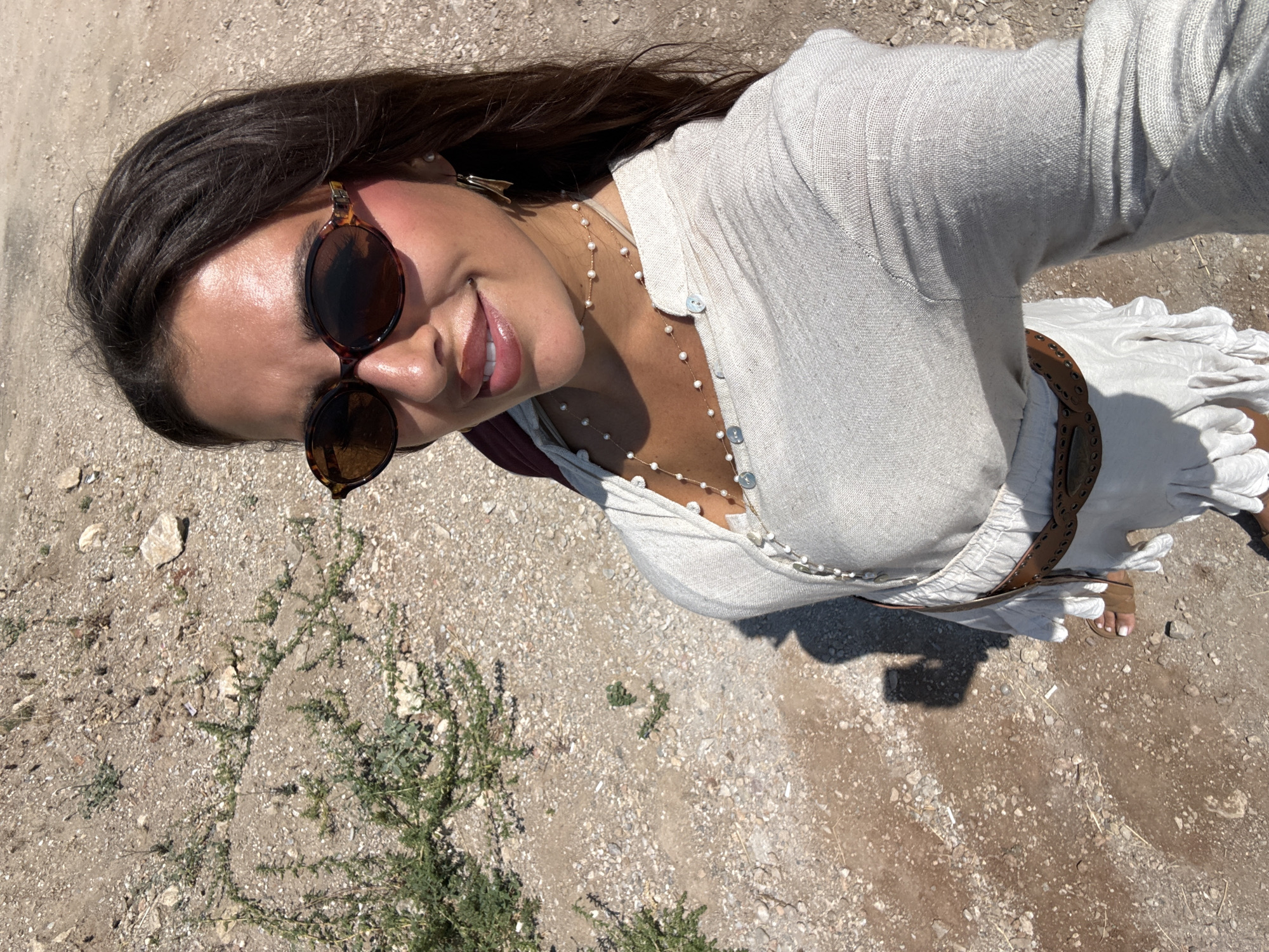 A woman is captured in a selfie outdoors, likely in Fethiye, Turkey. She is smiling and looking towards the camera, with her head tilted slightly. She has long, dark hair that falls over her shoulders and is wearing round, tortoiseshell sunglasses. A delicate pearl necklace adorns her neck, resting on a light-colored, long-sleeved linen shirt. The shirt has buttons down the front and appears to be layered over another white garment. A brown, woven belt cinches her waist. Her right foot, adorned with white toenail polish, is visible, wearing a brown sandal. The ground beneath her is composed of gravel, dirt, and small stones, with sparse green vegetation growing in patches. The lighting suggests it is daytime, with the sun casting shadows on the ground. The overall impression is one of a casual, sunny day. There is no visible text in the image.