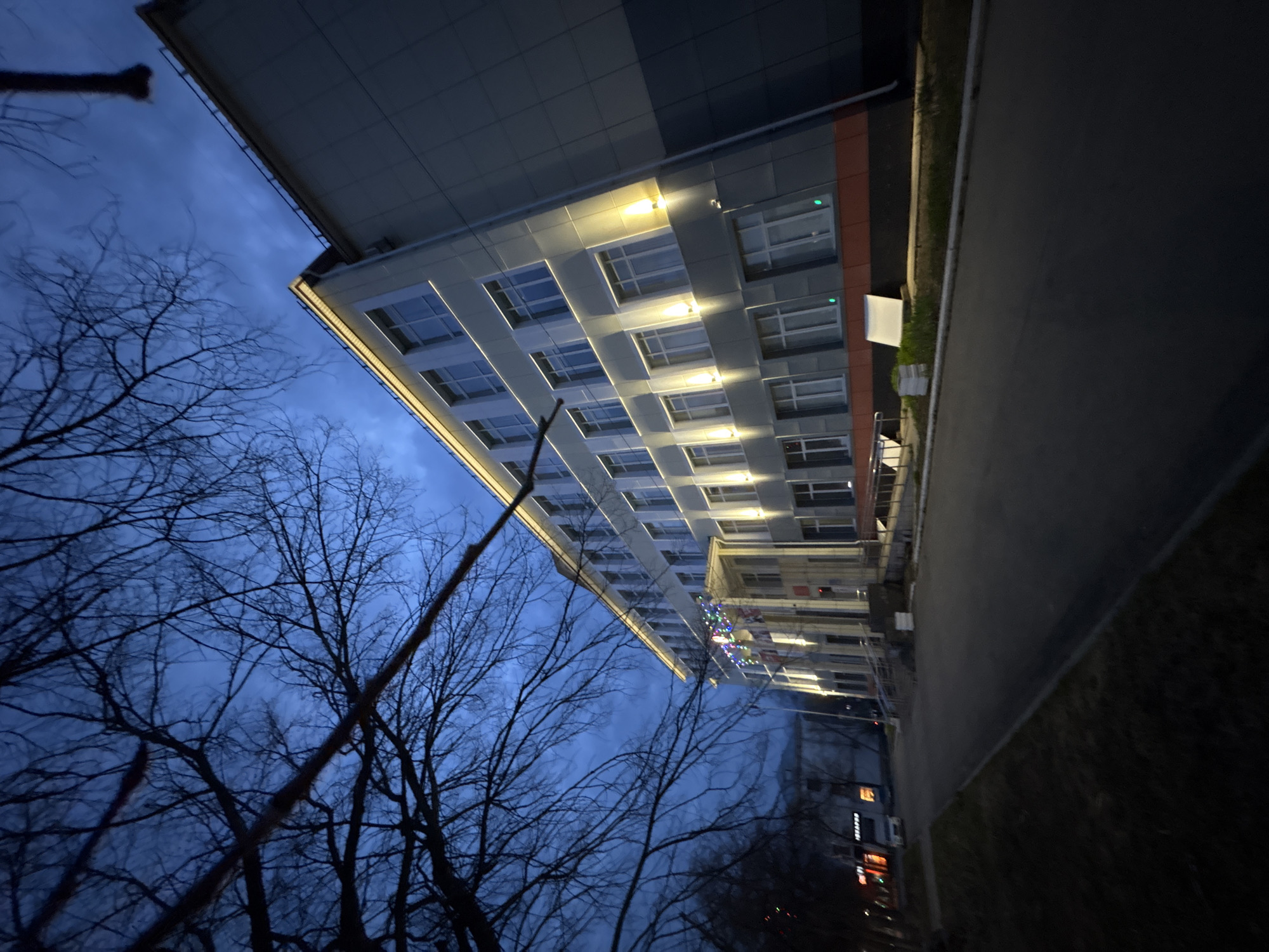 The image captures a modern multi-story building at night, viewed from a low angle, with bare trees silhouetted against the dark blue sky. The scene is illuminated primarily by artificial lights emanating from the building itself and some distant sources.
**Subjects:** The main subject is a contemporary building featuring a grey facade, possibly with sections of reddish-orange paneling on the right side. It has numerous rectangular windows, some of which appear to be lit from within. Prominent warm white or yellow LED strip lighting outlines the top edge of several floors, casting a bright glow on the exterior. To the left, a dense network of bare tree branches, devoid of leaves, occupies a significant portion of the frame, their dark forms contrasting sharply with the sky. A dark paved area, likely a road or sidewalk, runs along the right side of the building, bordered by a narrow strip of dark grass or foliage. In the distance along the building's facade, faint colored lights, possibly decorative, are visible. Other blurry, dark shapes and distant lights can be seen further down the street on the far right.
**Activity or event taking place:** No specific activity or event is depicted. The scene is static, capturing an urban or suburban environment during the late evening or night.
**Notable details:** The time of day is clearly night or deep twilight, as indicated by the dark sky and the reliance on artificial lighting. The sky is a deep, rich blue, suggesting some ambient light might still be present or a clear night sky. The bare trees strongly suggest a colder season, such as late autumn, winter, or early spring. The bright, deliberate lighting of the building creates a striking contrast with the natural darkness. The location, Chaikovsky, Russia, contributes to the cold-season atmosphere suggested by the trees.
**Visible text:** No legible text is visible anywhere in the image.