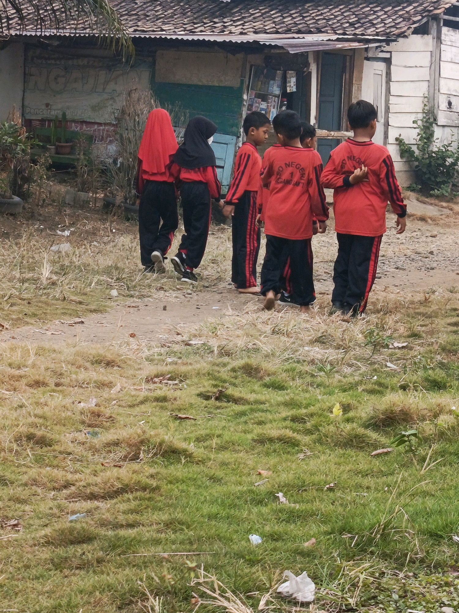 The image depicts five children, likely students based on their uniforms, walking away from the camera on a dirt path. They are in front of a modest, weathered building with a corrugated metal roof. The building appears to be a school or shop, with what looks like signage and possibly display shelves visible through a doorway. Two of the children on the left are wearing red headscarves, with one in black, and all are dressed in matching red long-sleeved shirts with black stripes on the sleeves and black pants with red stripes. The environment is somewhat unkempt, with dry grass, scattered leaves, and some visible debris, suggesting a rural or semi-urban setting. The children's attire and the building's condition hint at a location in a developing area, consistent with Megang Sakti II, Indonesia. The child second from the right has his hands clasped behind his back, while others walk with their hands by their sides.