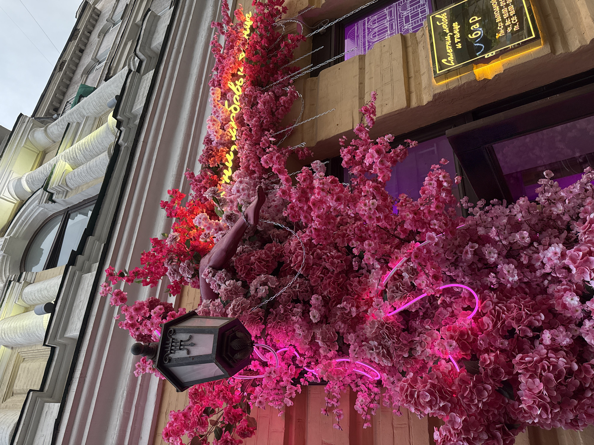The image captures a close-up view of a building facade adorned with abundant pink artificial flowers, possibly cherry blossoms. A vintage-style lantern is attached to the wall, draped with these flowers. A neon pink sign shaped like a heart is partially visible amidst the floral display. Further to the right, a yellow illuminated sign with Russian text is mounted on the brick wall. The sign reads "Свет, любовь и танцы" (Light, love, and dances) above the word "бар" (bar), followed by opening hours and a website address. The architectural style suggests an older building, with decorative elements and arched windows visible in the background. The overall impression is one of vibrant decoration, likely for a bar or establishment aiming for a romantic or festive ambiance. The presence of the Russian text on the sign definitively places this scene in Russia, and the specific text on the sign indicates it is likely a bar named "Свет, любовь и танцы" in Krasnoyarsk, Russia.