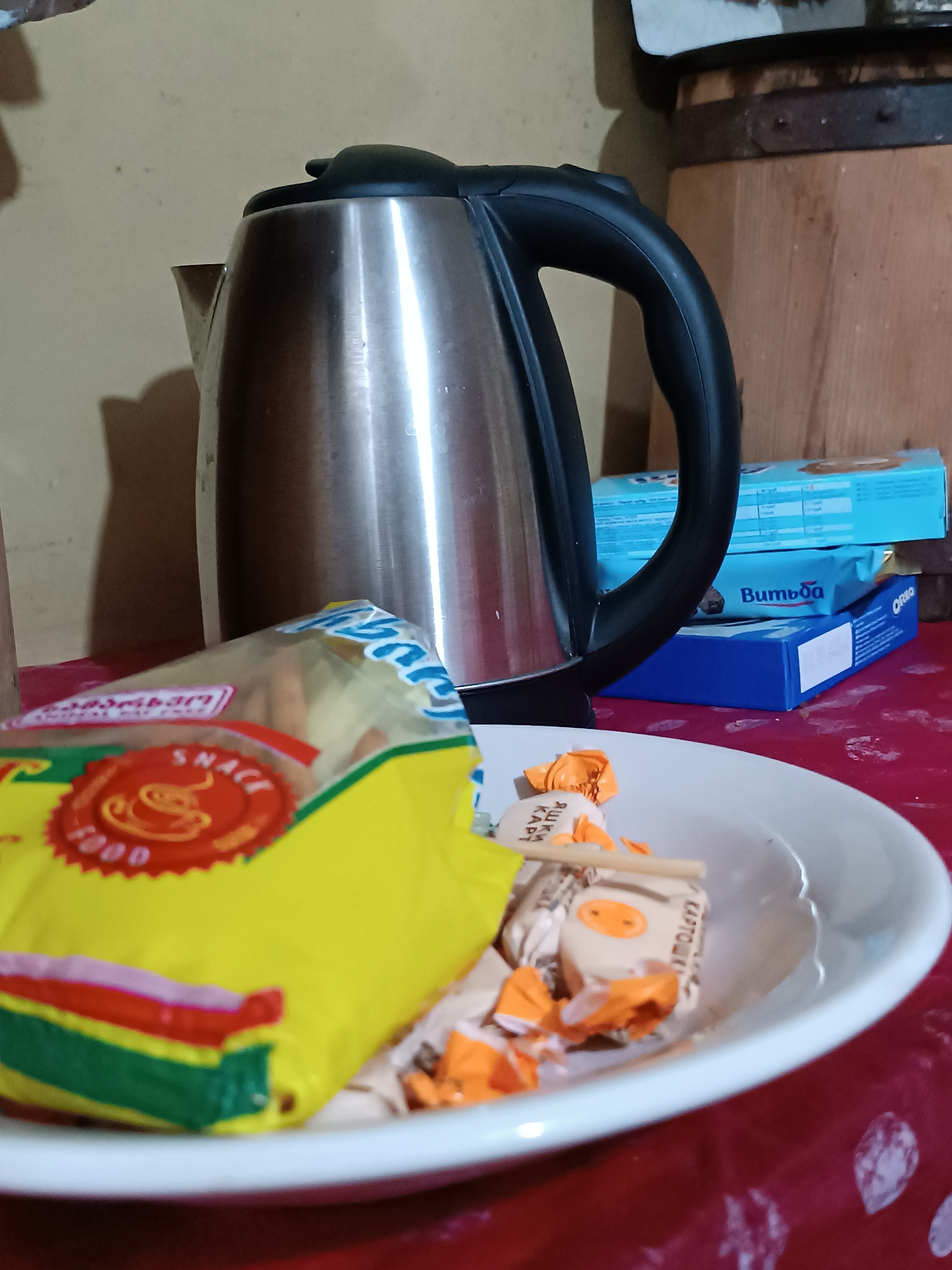 The image depicts a still life on a red tablecloth. In the foreground, a white ceramic bowl contains several individually wrapped candies, some with orange wrappers and others with cream-colored wrappers featuring orange cartoon faces and text. A wooden chopstick rests across the bowl. To the left of the bowl is a yellow snack bag with colorful stripes along the bottom edge, and a circular red emblem with text and an icon of a steaming cup. In the background, a stainless steel electric kettle with a black handle is prominent. Behind the kettle, stacked on top of each other, are three blue boxes. The top box is partially obscured, the middle box has the word "Вимбоа" (Vimbaa) printed on it, and the bottom box has the word "Oreo" printed on it. To the far right, a portion of a wooden barrel with metal bands is visible. The background wall is a light, neutral color. The scene appears to be set indoors, possibly in a kitchen or dining area, with artificial lighting casting shadows. There are no people visible in the image. The objects suggest a setting where snacks and beverages are prepared or consumed. There is no visual information within the image that provides any specific details about the location being in Khashuri, Georgia.