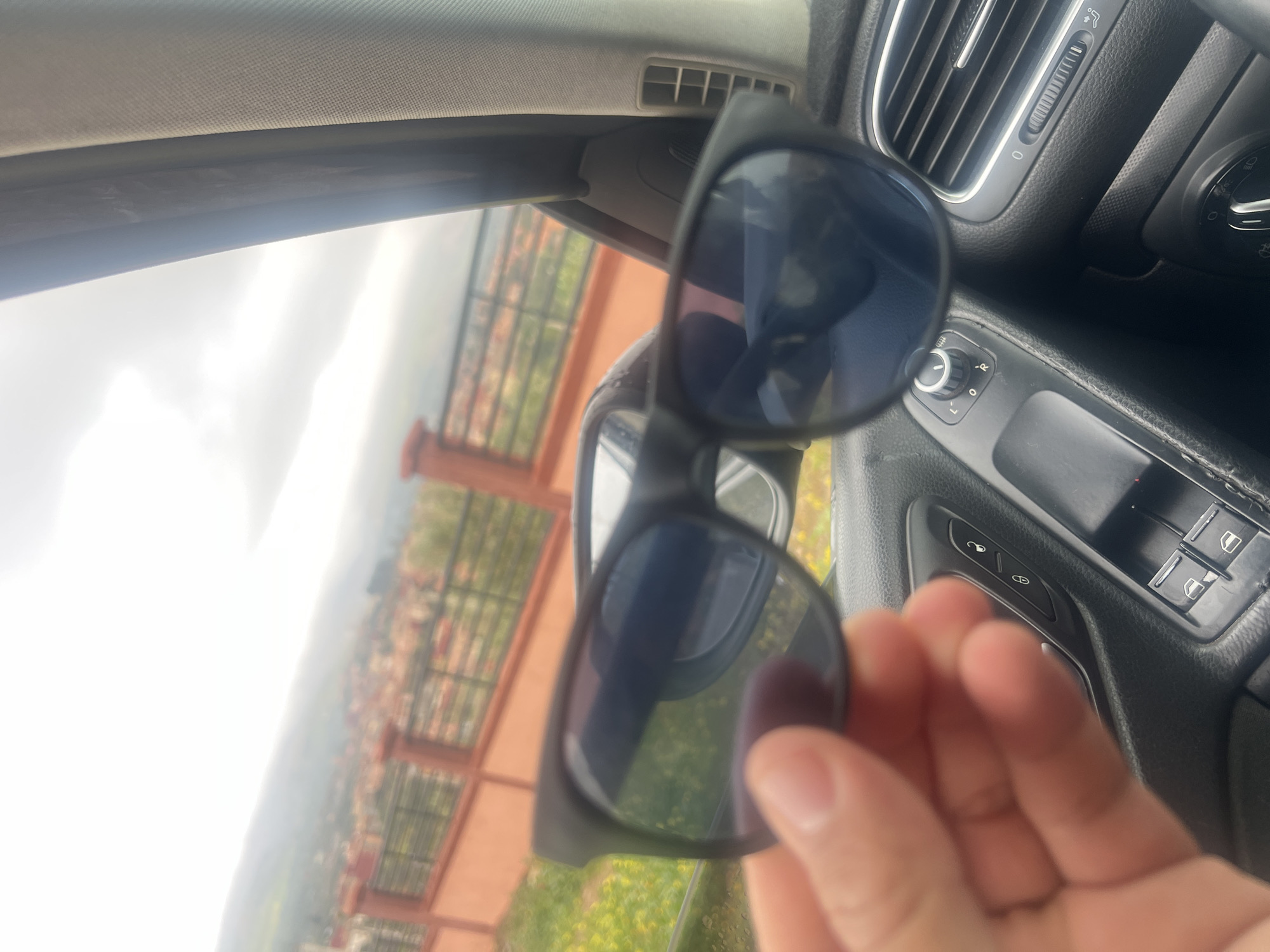 A pair of black-framed sunglasses are held up inside a car, partially obscuring a view of Ouaouizert, Morocco. The car's dashboard and door panel are visible, with controls for air vents, mirrors, and windows. Outside, a landscape of buildings and greenery is seen through a fence, under a cloudy sky. A hand partially grips the sunglasses.