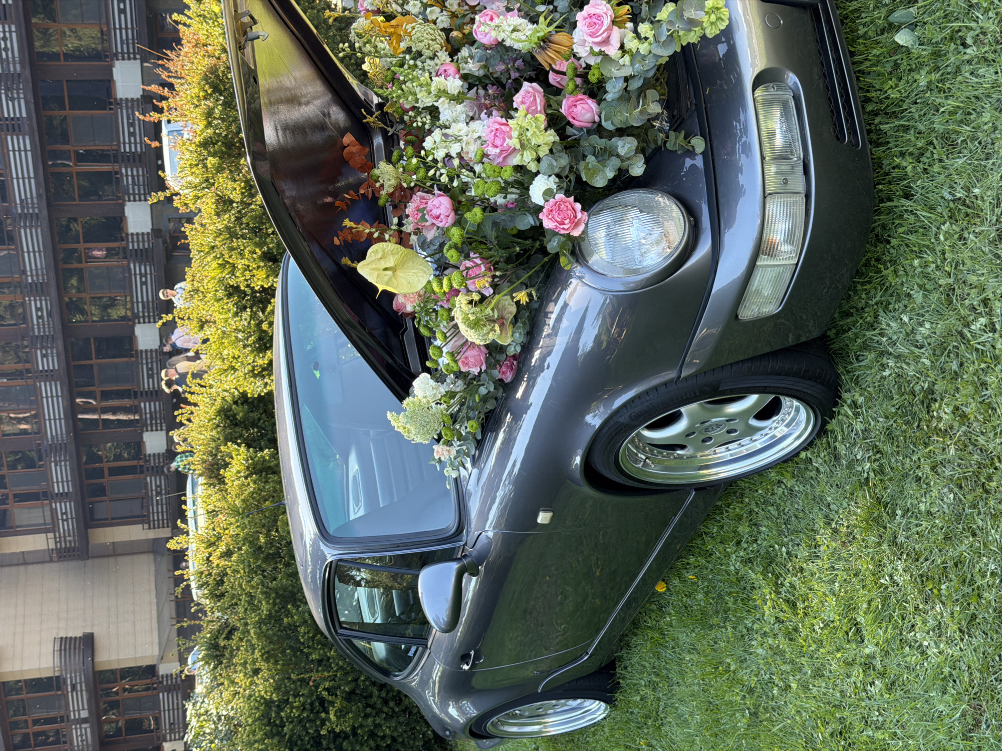 A gray Porsche 911 stands on a green lawn adjacent to a tree-lined building. The hood is open, overflowing with a colorful assortment of flowers, including pink roses and green foliage. People are visible in the background. The location is likely Repino, Russia.
