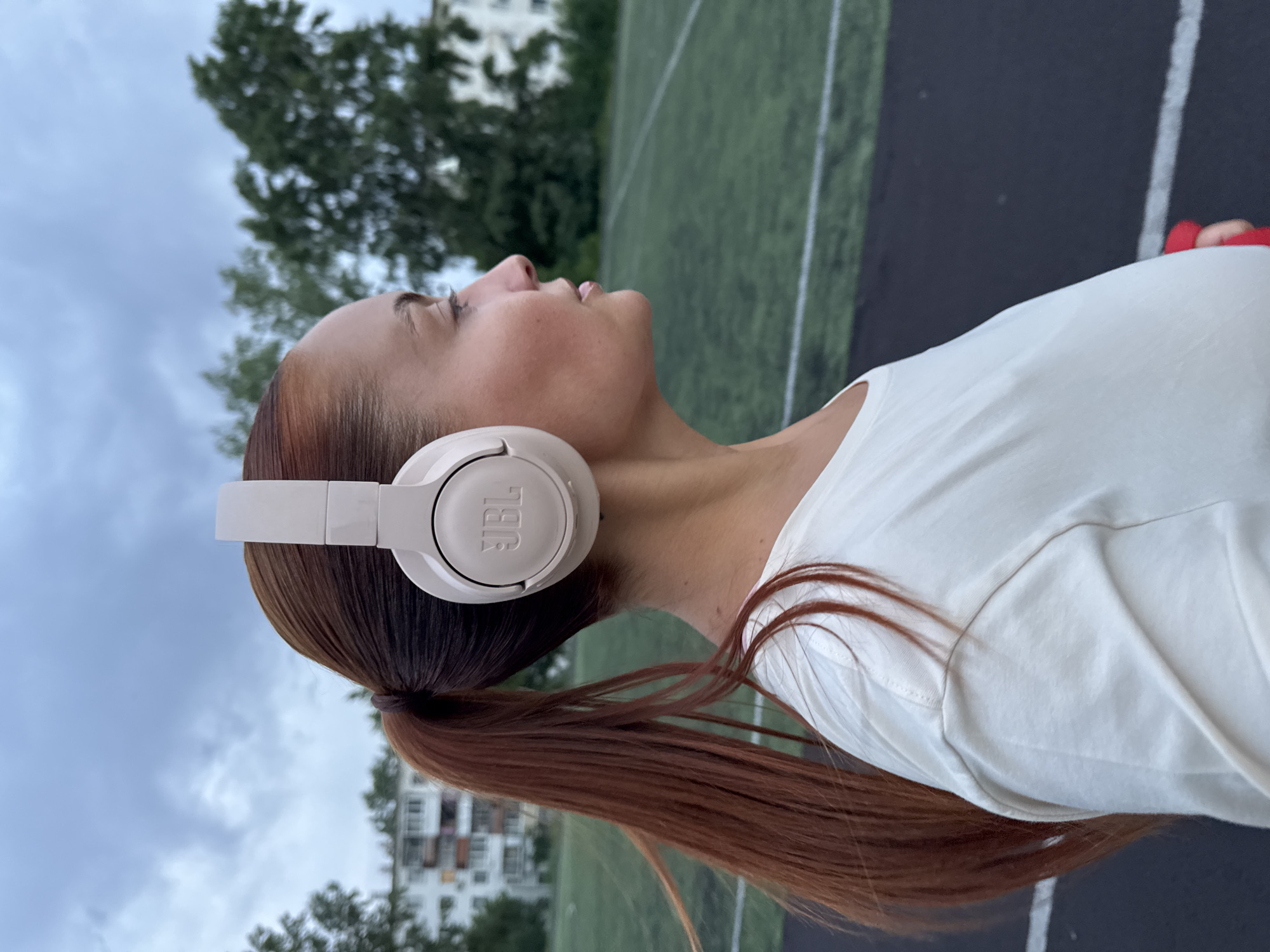 A young woman wearing a white t-shirt and beige JBL headphones stands outdoors. Her auburn hair is pulled back in a ponytail. She is looking up and to the right. The background features a running track with green turf and black asphalt lanes with white lines. In the distance are trees and a white multi-story building, indicating an urban environment. This is the city of Chaikovsky, Russia.