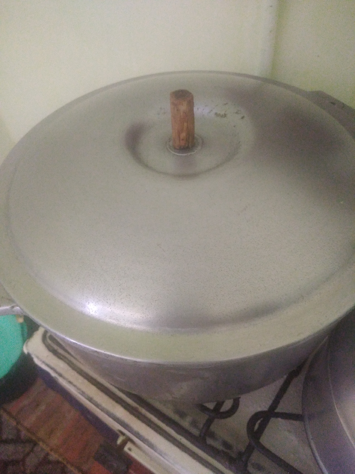 A close-up, medium-angle shot shows a silver pot with a wooden handle lid on a stove. The pot is circular and has a rounded shape. The lid is also circular and has a small, cylindrical wooden knob in the center. The pot is sitting on what appears to be a stovetop, with some cooking elements visible underneath. To the left of the pot, a portion of a green bowl or container is visible. The background is a light-colored wall. The lighting in the image is somewhat dim, creating soft shadows. The overall scene suggests a domestic setting, likely a kitchen.