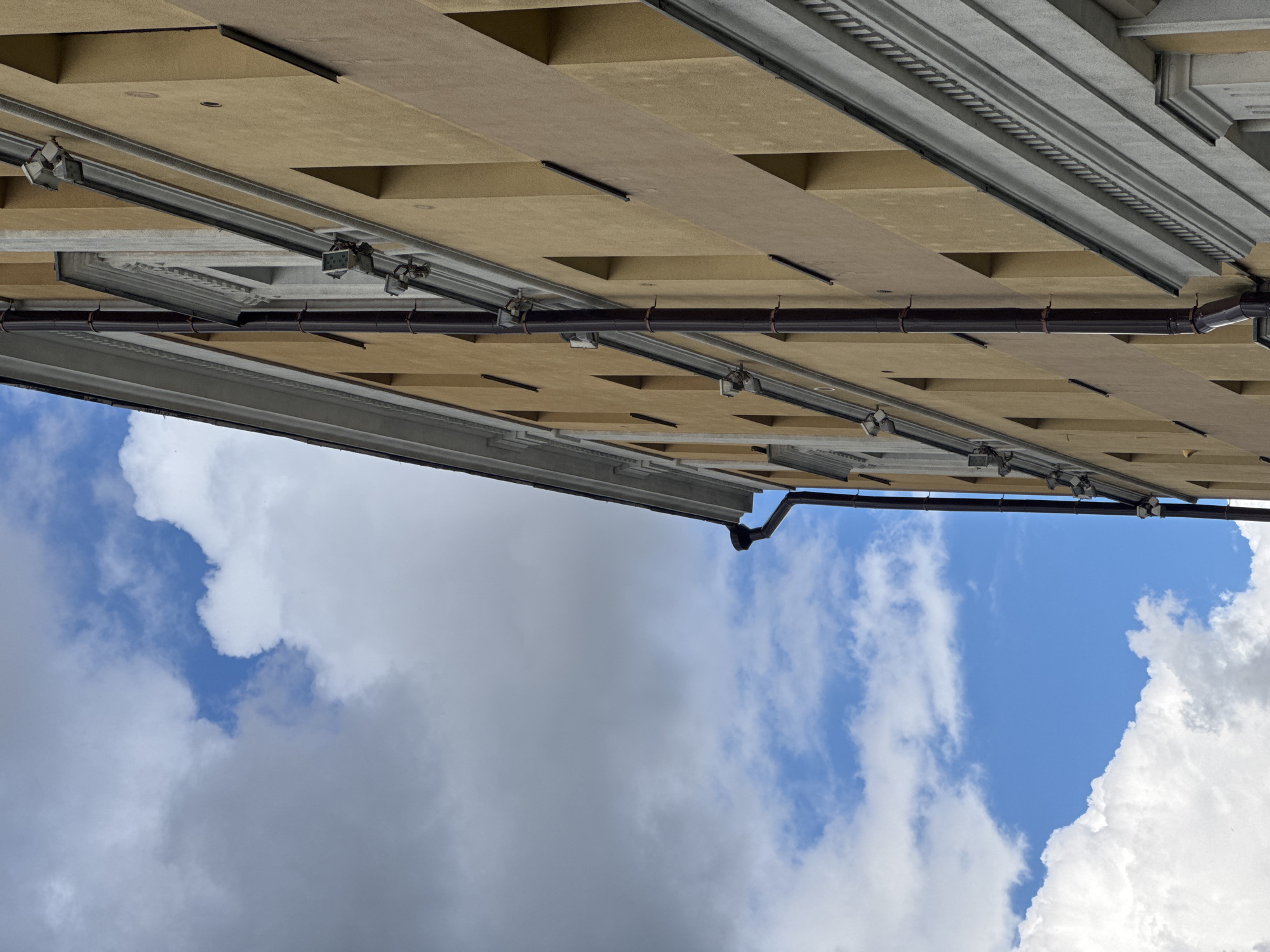 The media file shows an extreme upward angle view of a building's facade and the sky above. The building appears to be made of light brown rectangular panels with a grid-like pattern. Silver tracks and brown pipes run along the underside of the building's overhang, with several small, silver fixtures attached to the tracks. The sky is a mix of bright blue and white clouds, with some areas of darker, more diffused clouds suggesting a dynamic weather pattern. The lighting suggests it is daytime, likely midday or afternoon, given the brightness of the sky. There is no visible text in the image. The perspective creates a sense of looking up into the sky from a very low vantage point, possibly at the base of a tall building. The architectural details of the building, such as the metallic tracks and fixtures, suggest a modern or recently constructed building. The presence of clouds indicates it is not a perfectly clear day, but the patches of blue suggest it is not heavily overcast. Without further context, specific location cues beyond Moscow, Russia are not evident. The image focuses on architectural elements and the sky, creating a rather abstract composition.
